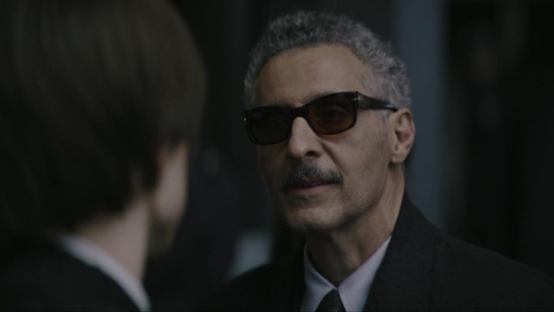 A man with short hair and sunglasses in a suit speaks to another person facing away