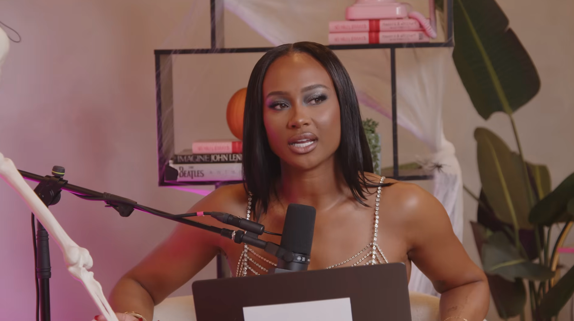 Person in elegant dress speaking into a microphone during a podcast recording, surrounded by books and plants