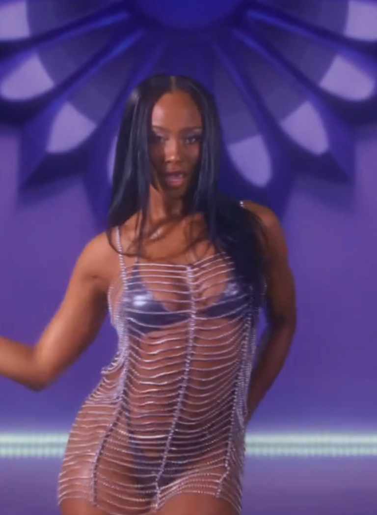 A person poses in a netted dress over a metallic outfit, set against a geometric backdrop