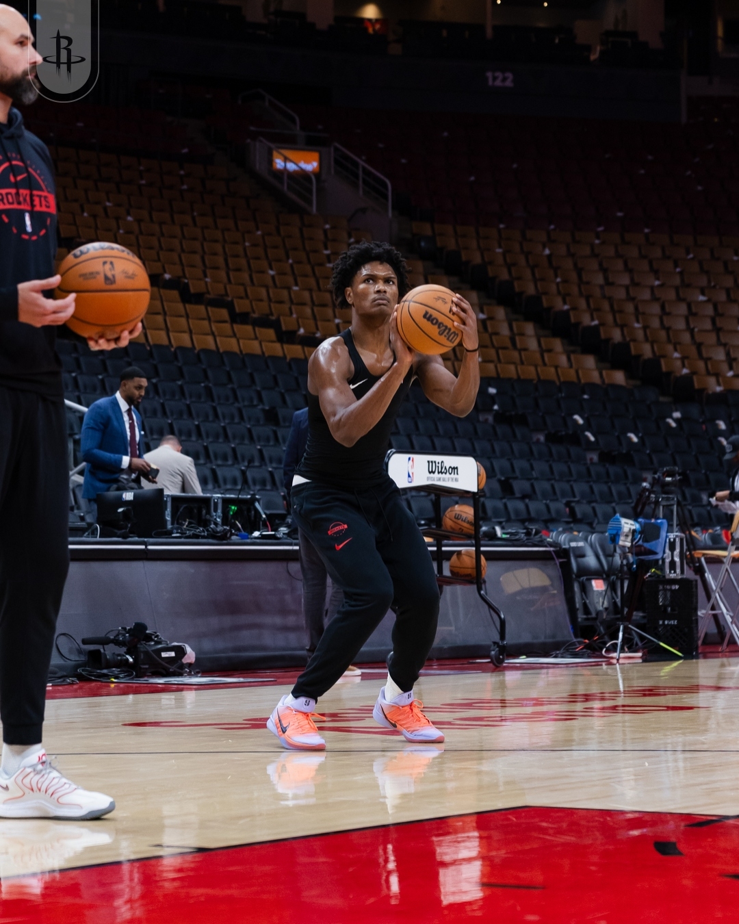 Basketball player practicing shooting on a court, wearing athletic attire and sneakers, with an empty arena in the background