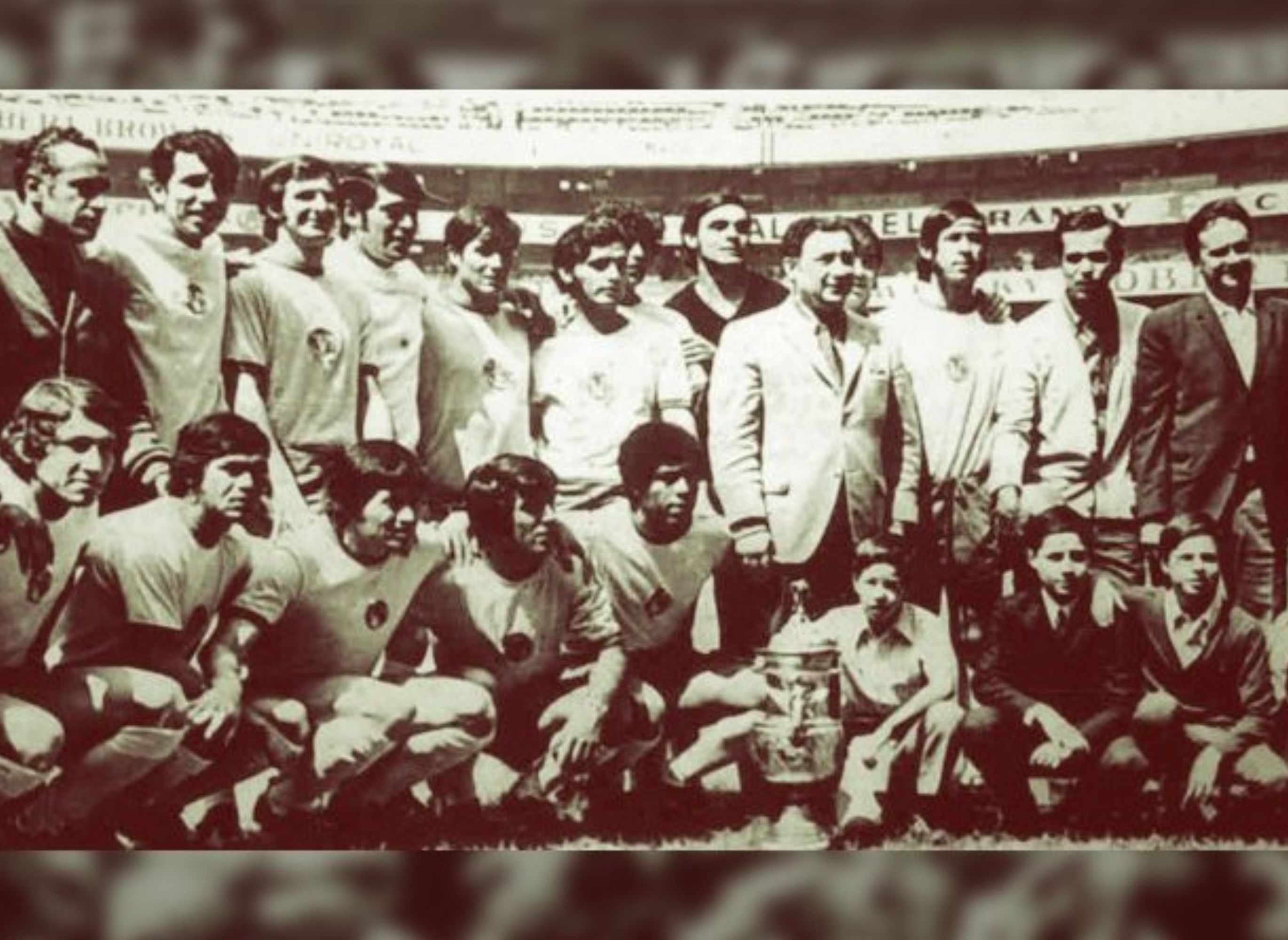 A vintage photo of a soccer team and staff posing in a stadium. The group includes players in uniforms and others in suits