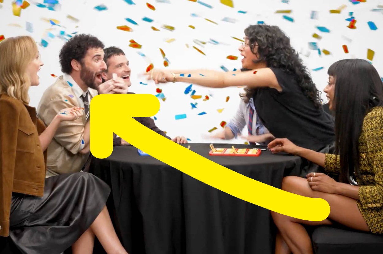 Group of people at a table playing a game, with one person excitedly pointing while others watch, surrounded by confetti