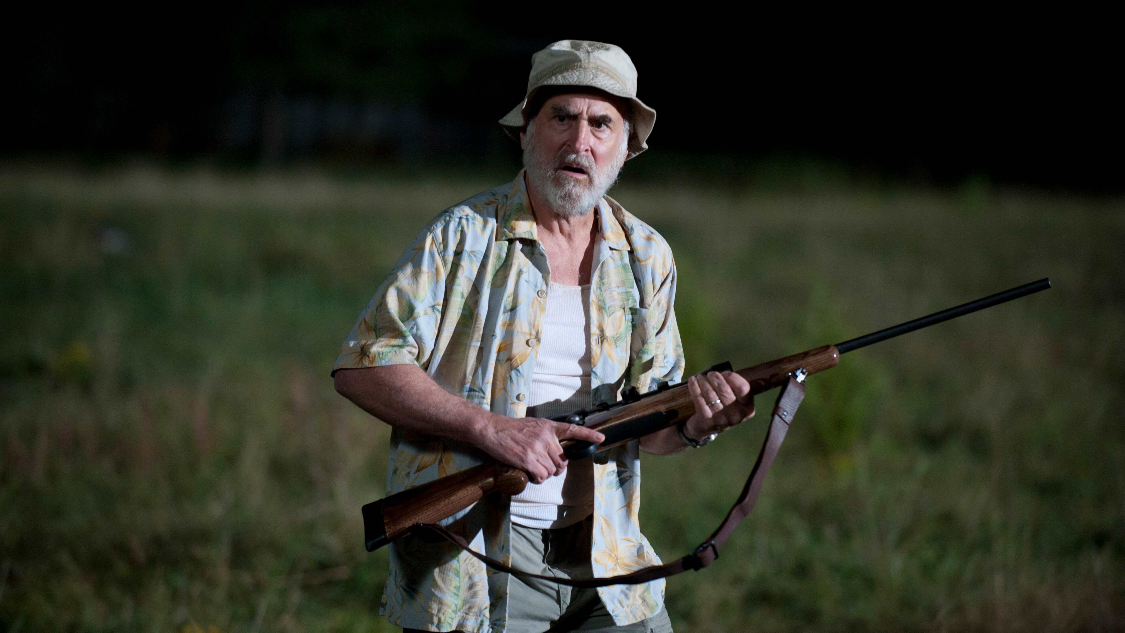 An older antheral   with a beard wears a hat, tropical shirt, and holds a rifle, lasting  successful  a grassy field, looking amazed  oregon  alert