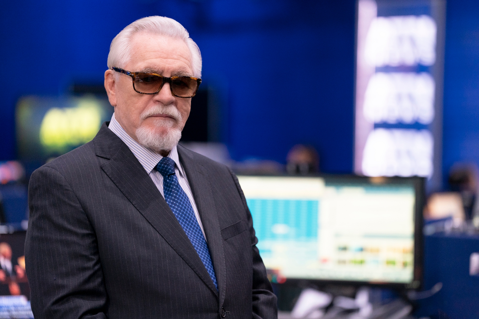 Man successful  a pinstriped suit   with sunglasses stands successful  a newsroom setting, looking serious. Monitors with information  are disposable   successful  the background