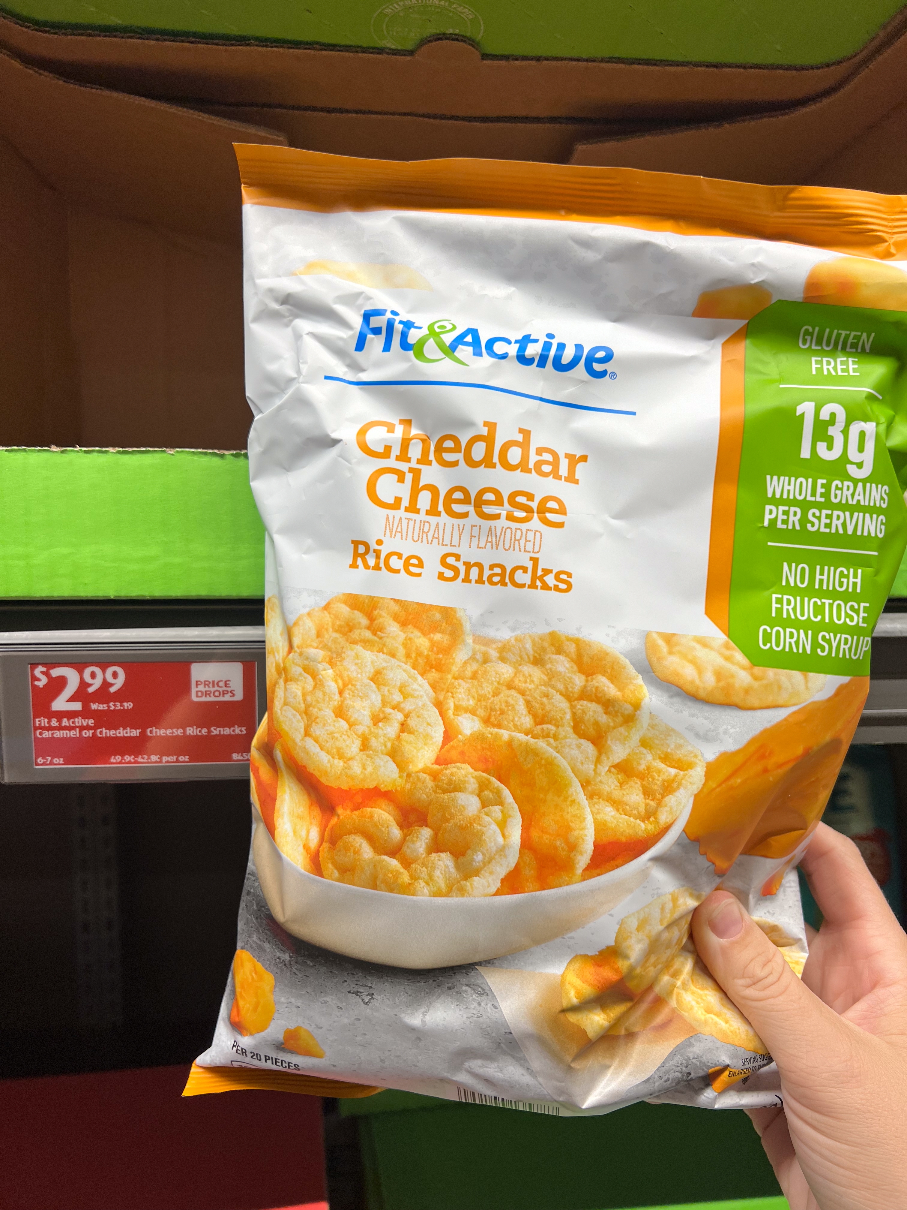 Person holding a bag of Fit &amp;amp; Active Cheddar Cheese Rice Snacks priced at $2.99. Packaging highlights gluten-free and natural flavor features
