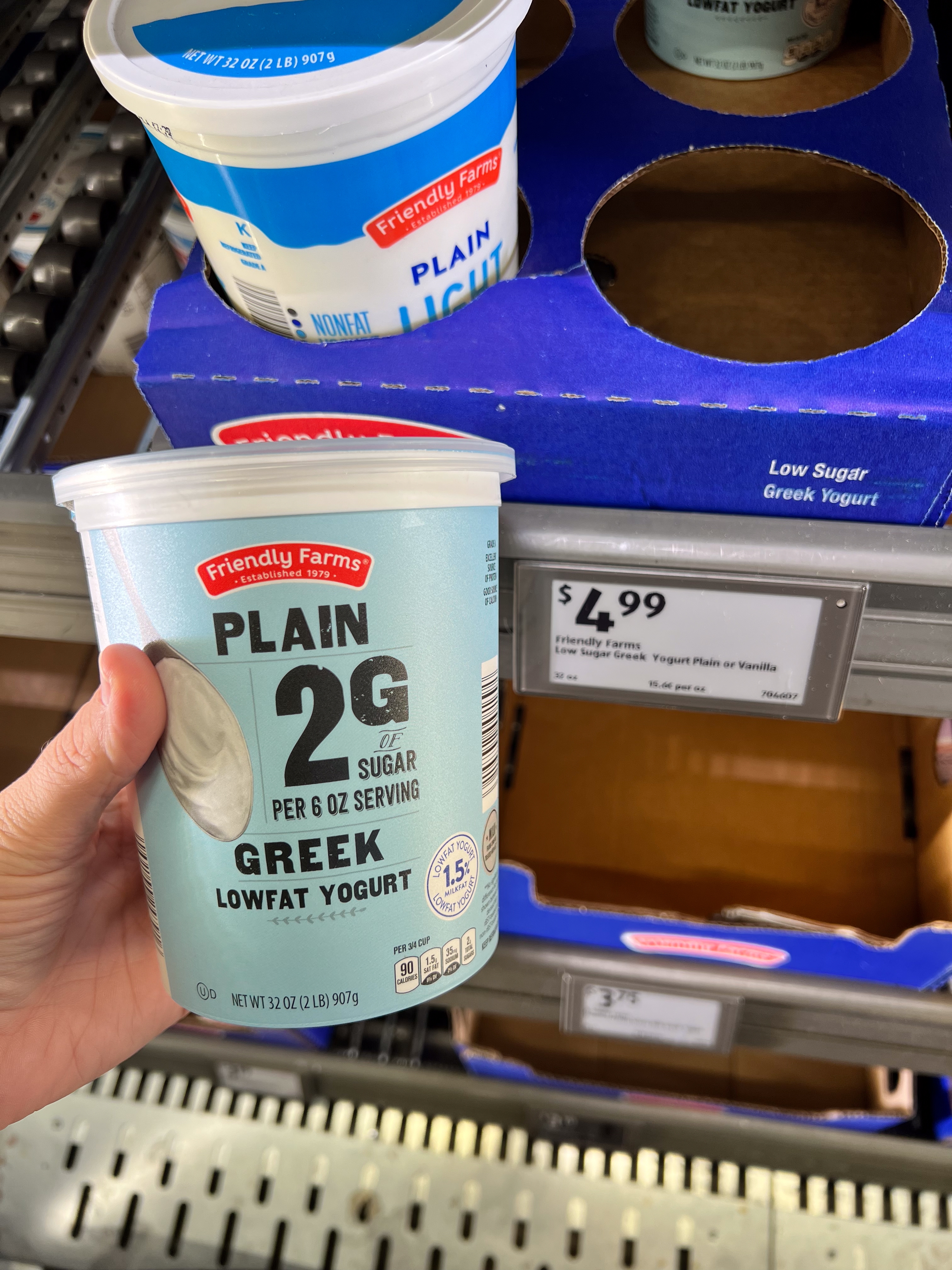 A person is holding a tub of Friendly Farms Plain Greek low-fat yogurt with 2g of sugar, priced at $4.99 on the shelf