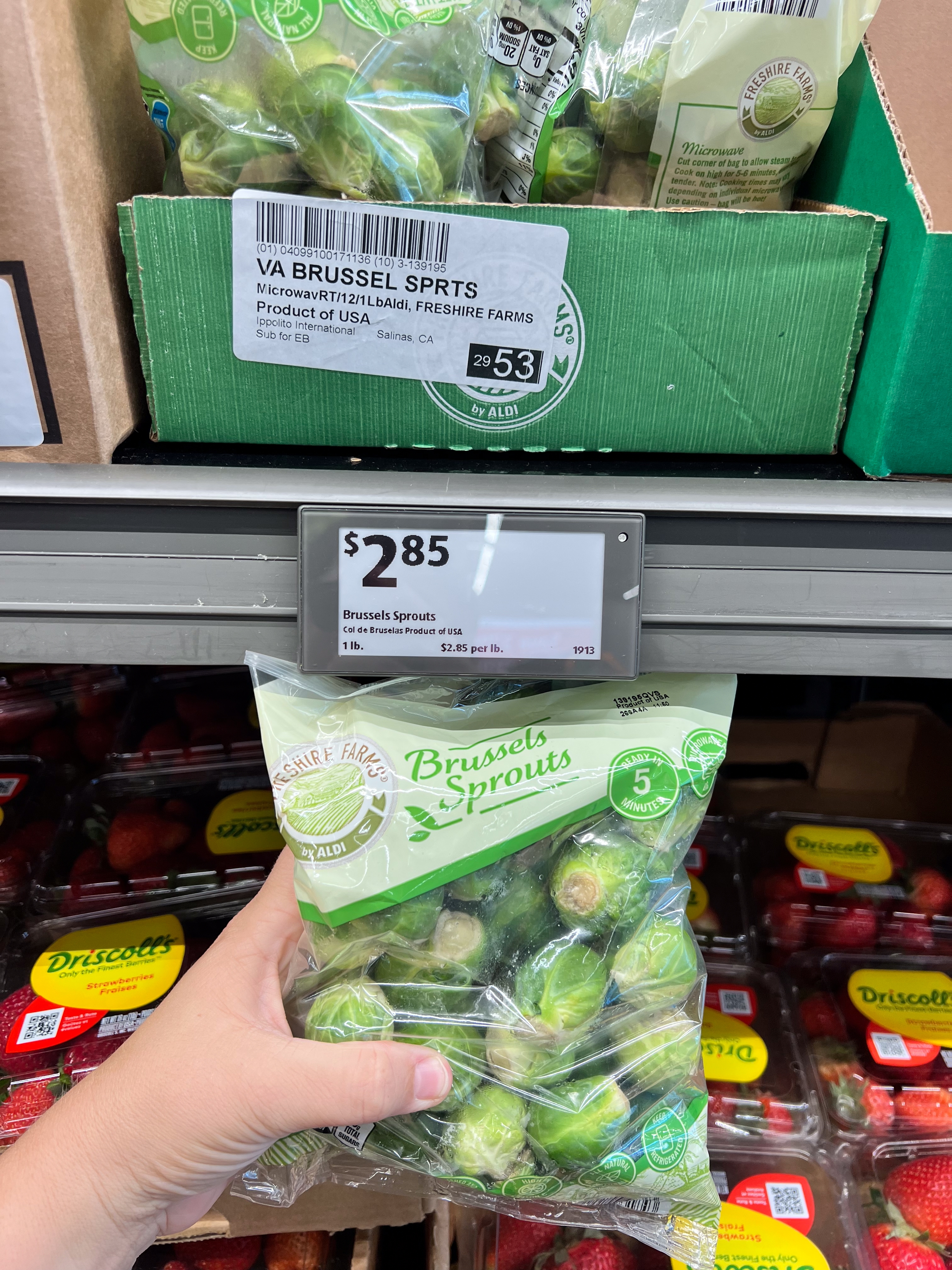 Hand holding a bag of Brussels sprouts near a store shelf, labeled $2.85 per package. Other produce items are visible in the background