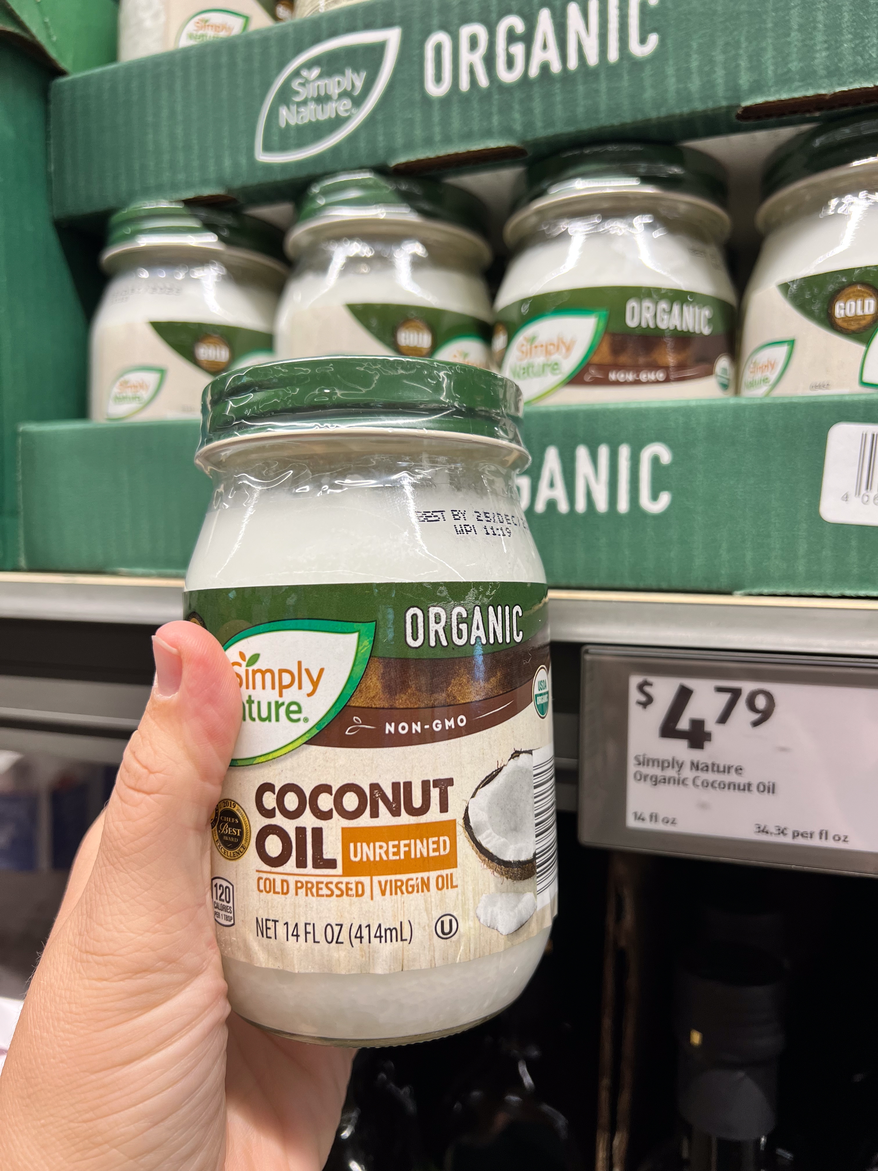 Hand holding a jar of Simply Nature Organic Unrefined Coconut Oil in a store. Price displayed: $4.79