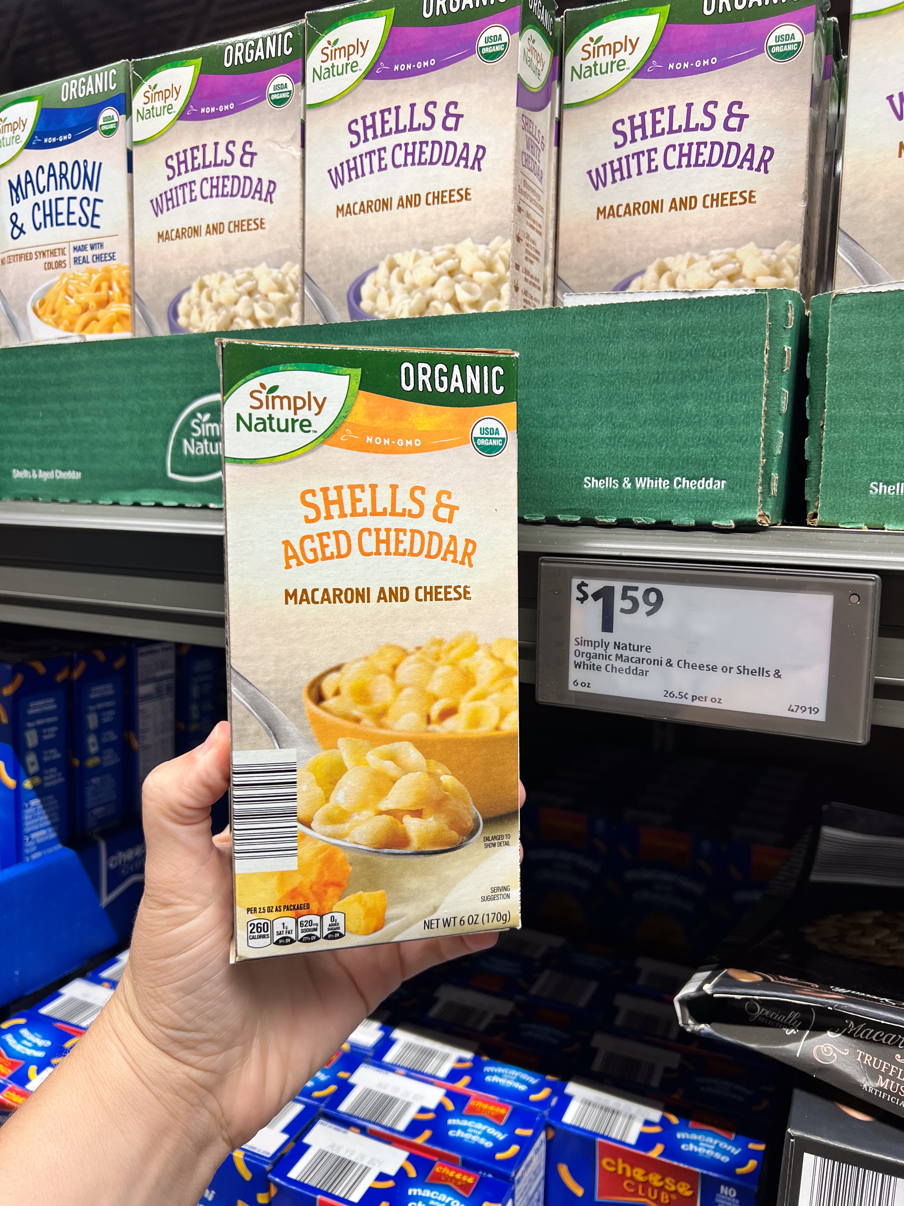 Person holding a box of Simply Nature organic shells and aged cheddar macaroni and cheese in a store. Price tag reads $1.59