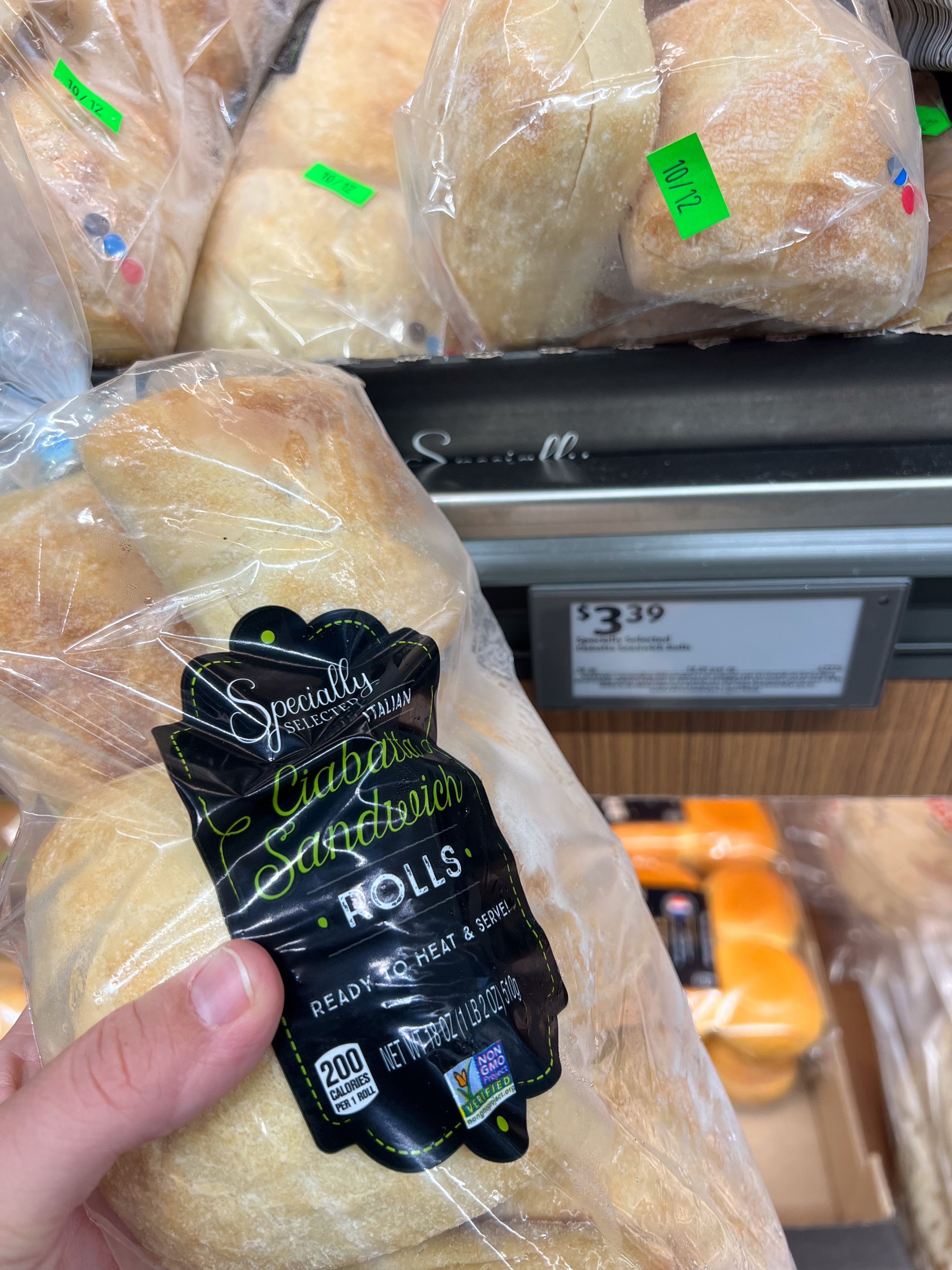 A hand holds a package of sandwich ciabatta rolls in a grocery store. Price tag reads $3.39
