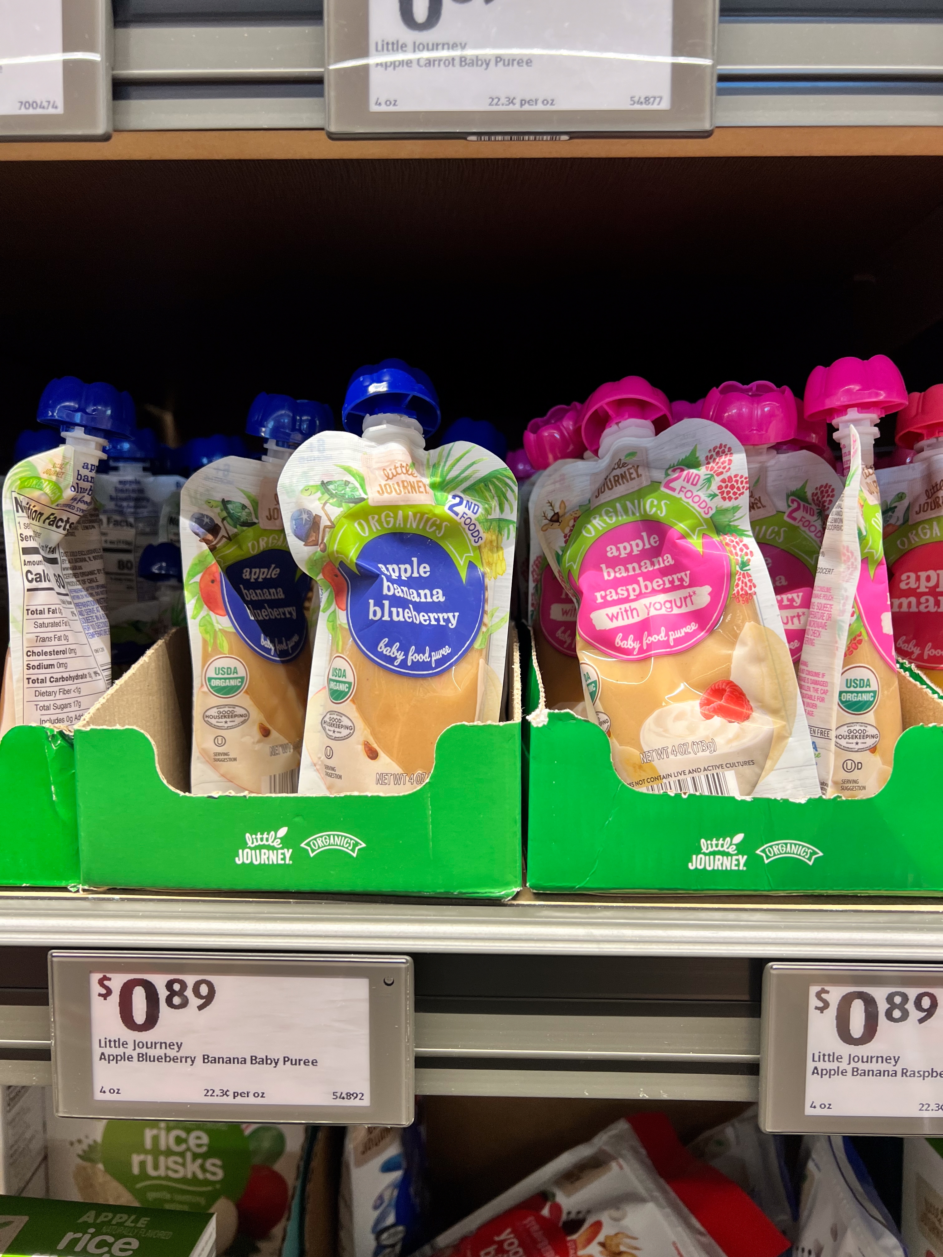 A grocery store shelf displays baby food pouches in apple banana blueberry and apple banana raspberry flavors, priced at $0.89 each