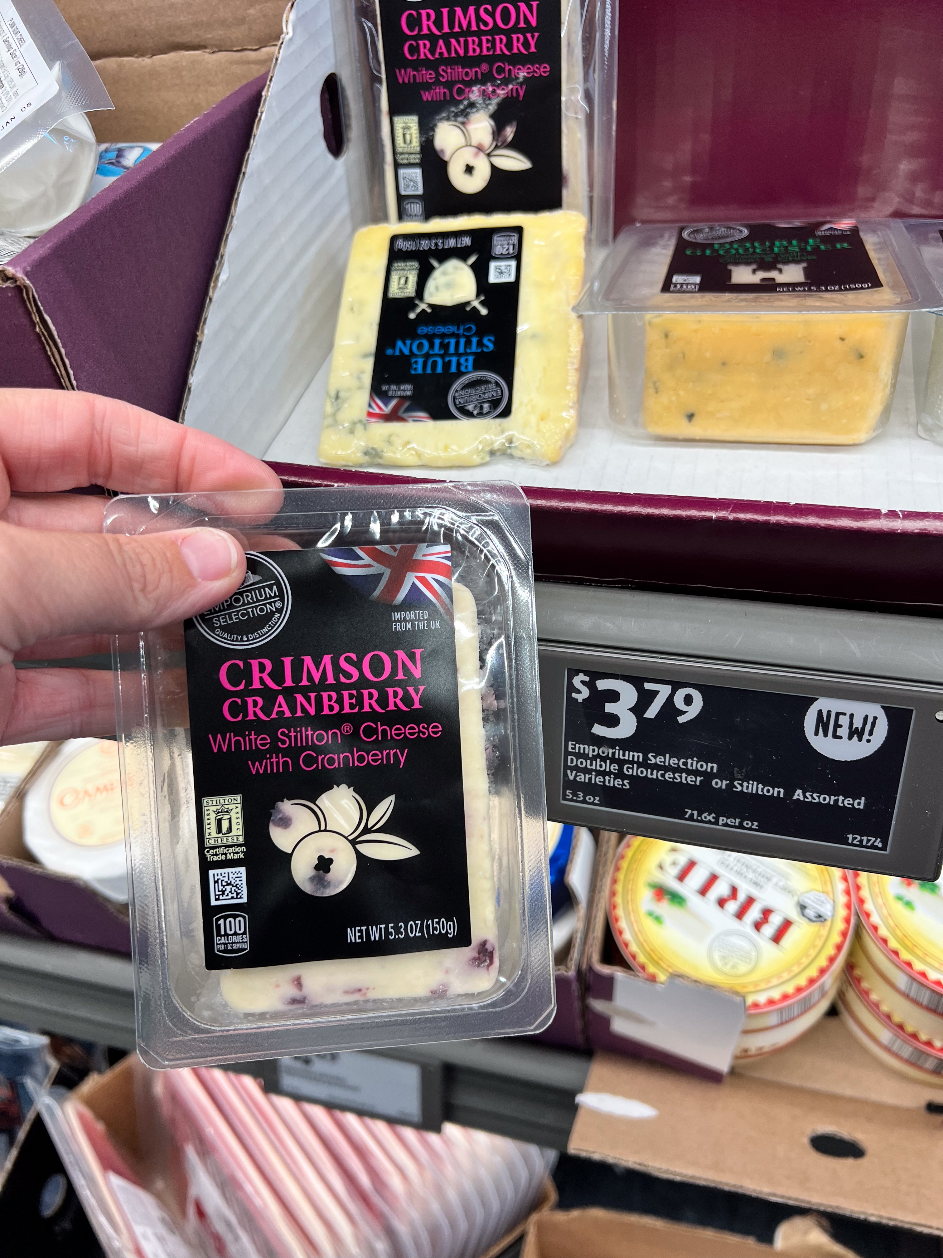 Person holding packaged white Stilton cheese with cranberries. Price tag reads $3.79 for assorted cheeses. Several cheese varieties visible in store display