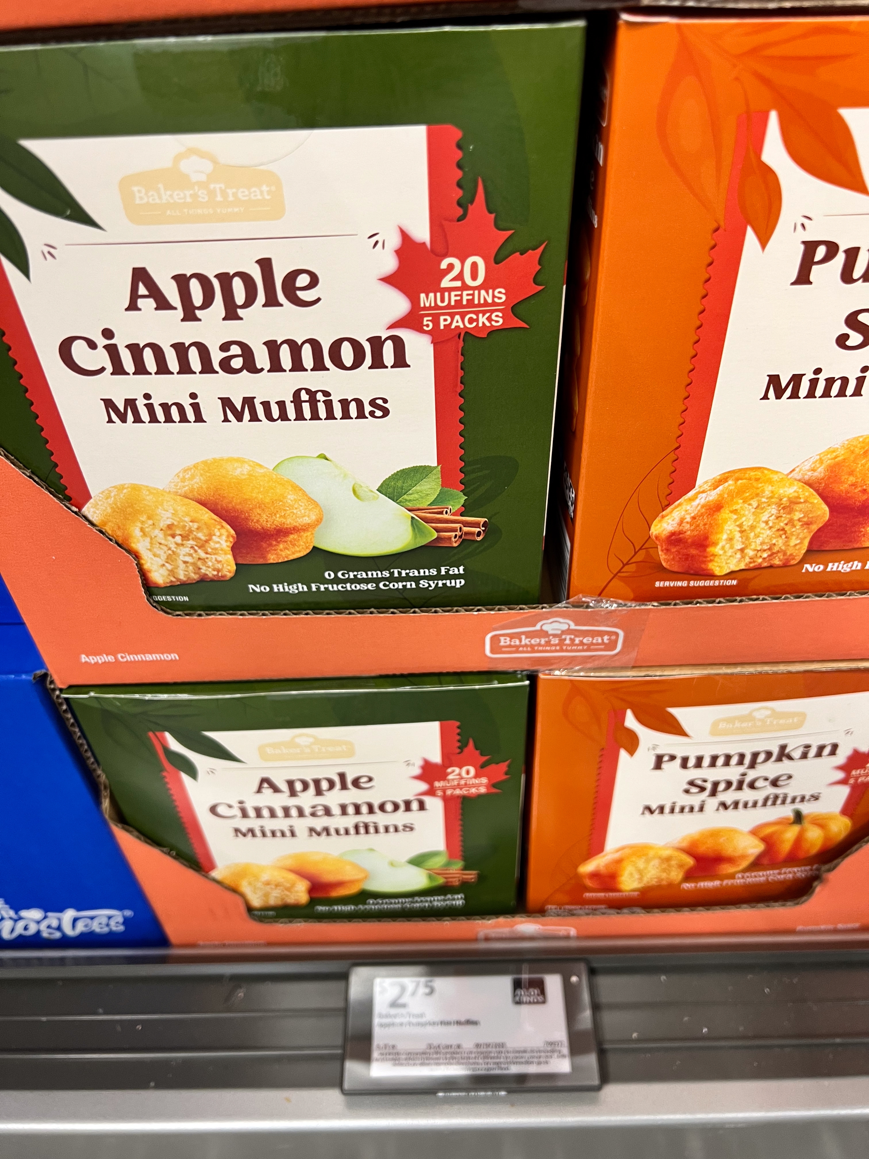 Apple Cinnamon and Pumpkin Spice mini muffins boxes on a store shelf, promoting 20 muffins in 5 packs. Sign shows pricing details