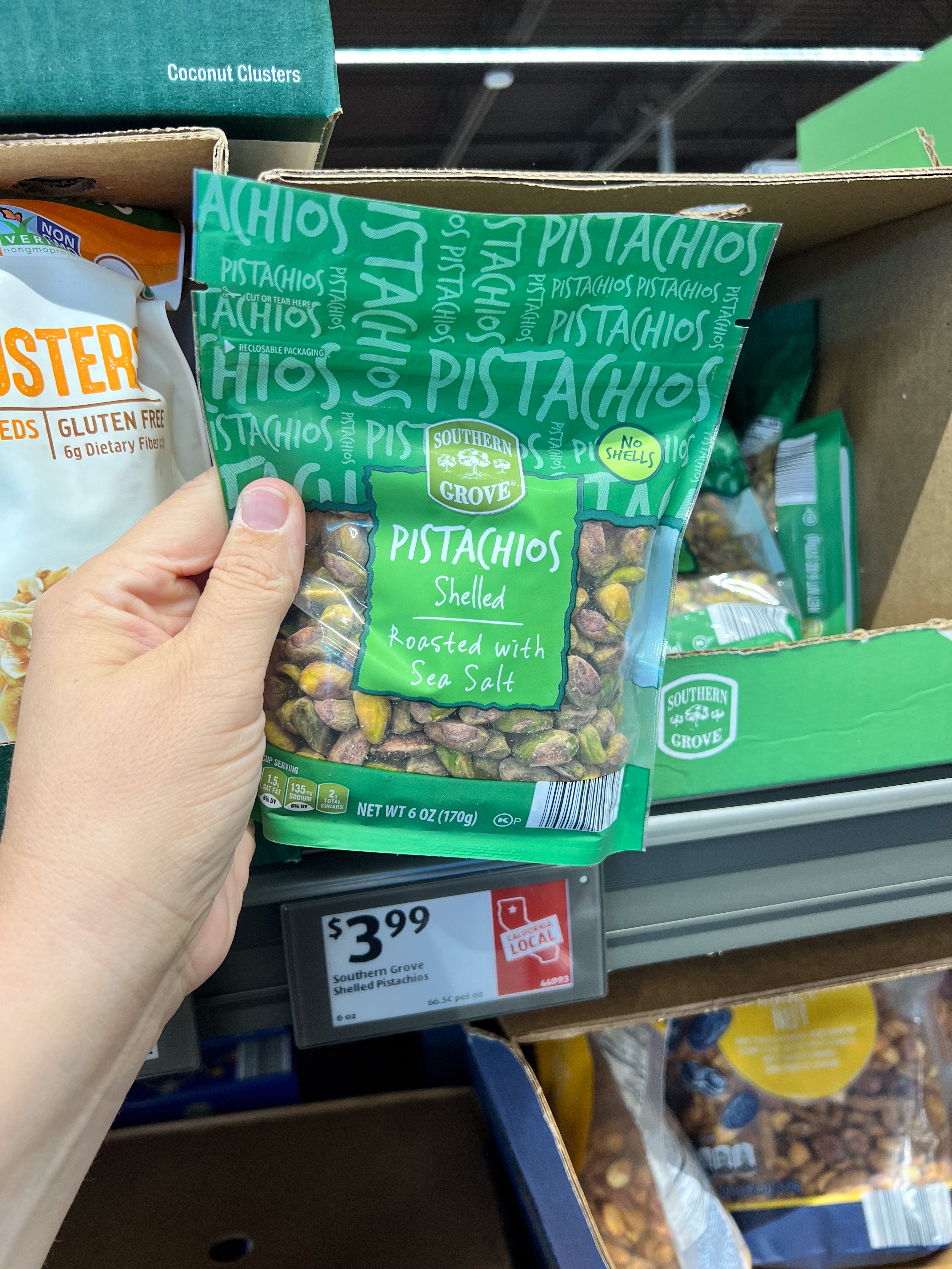 Bag of Southern Grove pistachios, roasted with sea salt, held by a person in a store aisle. Price tag shows $3.99
