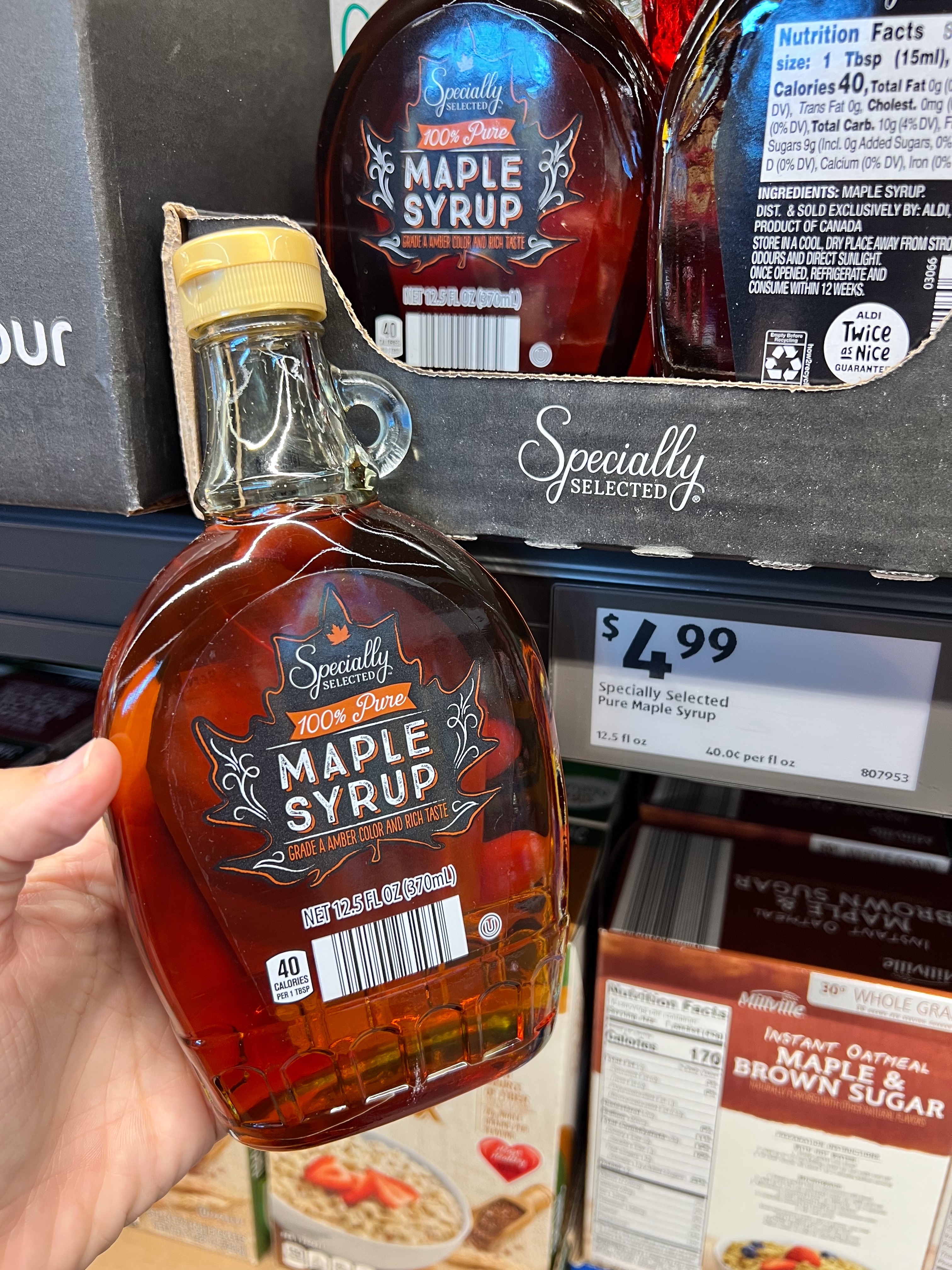 Person holding a bottle of Specially Selected 100% Pure Maple Syrup in a store, priced at $4.99