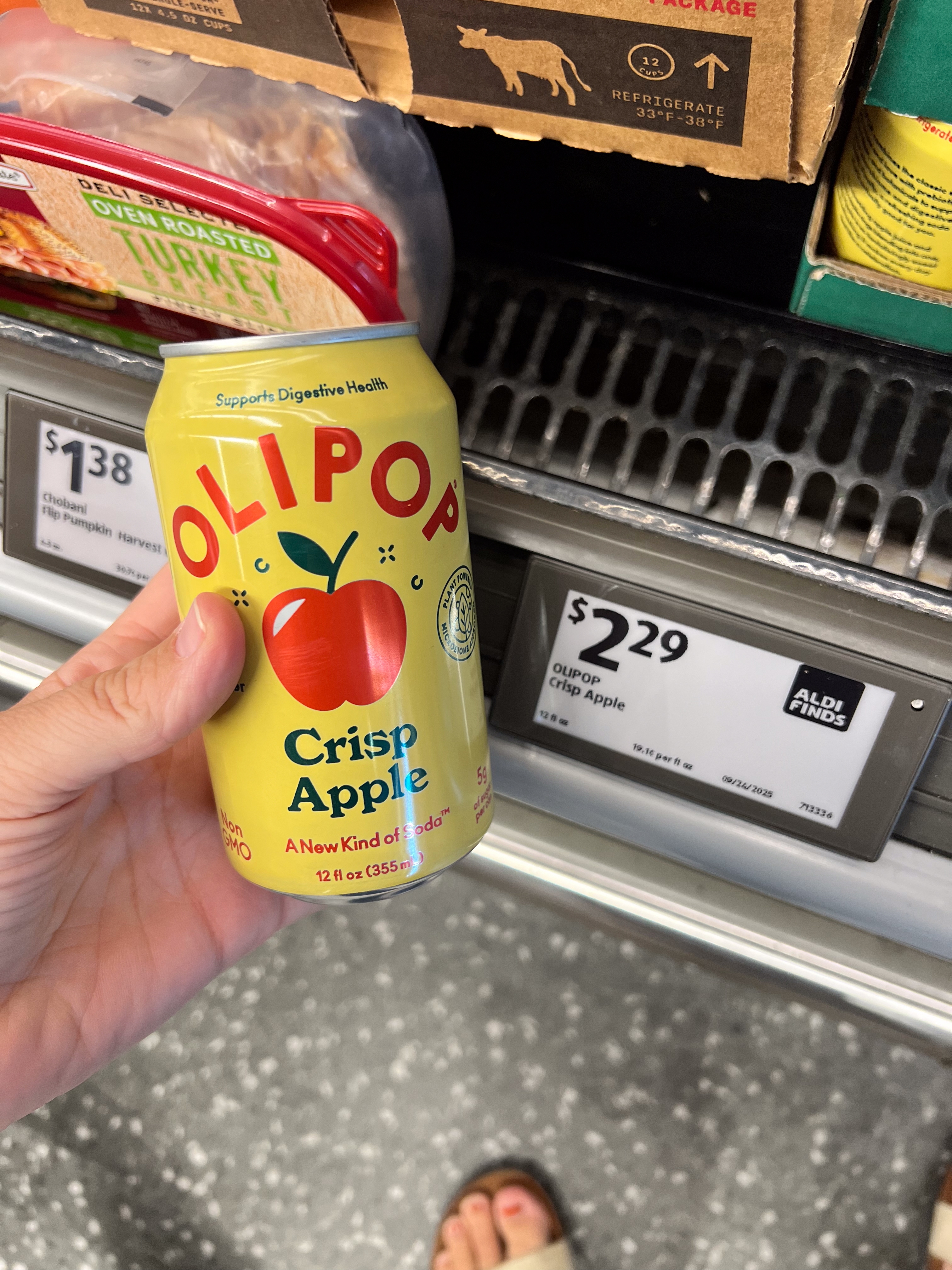 Hand holding Olipop Crisp Apple soda can, on a grocery store shelf. Price tag shows $2.29. Nearby are packages of turkey