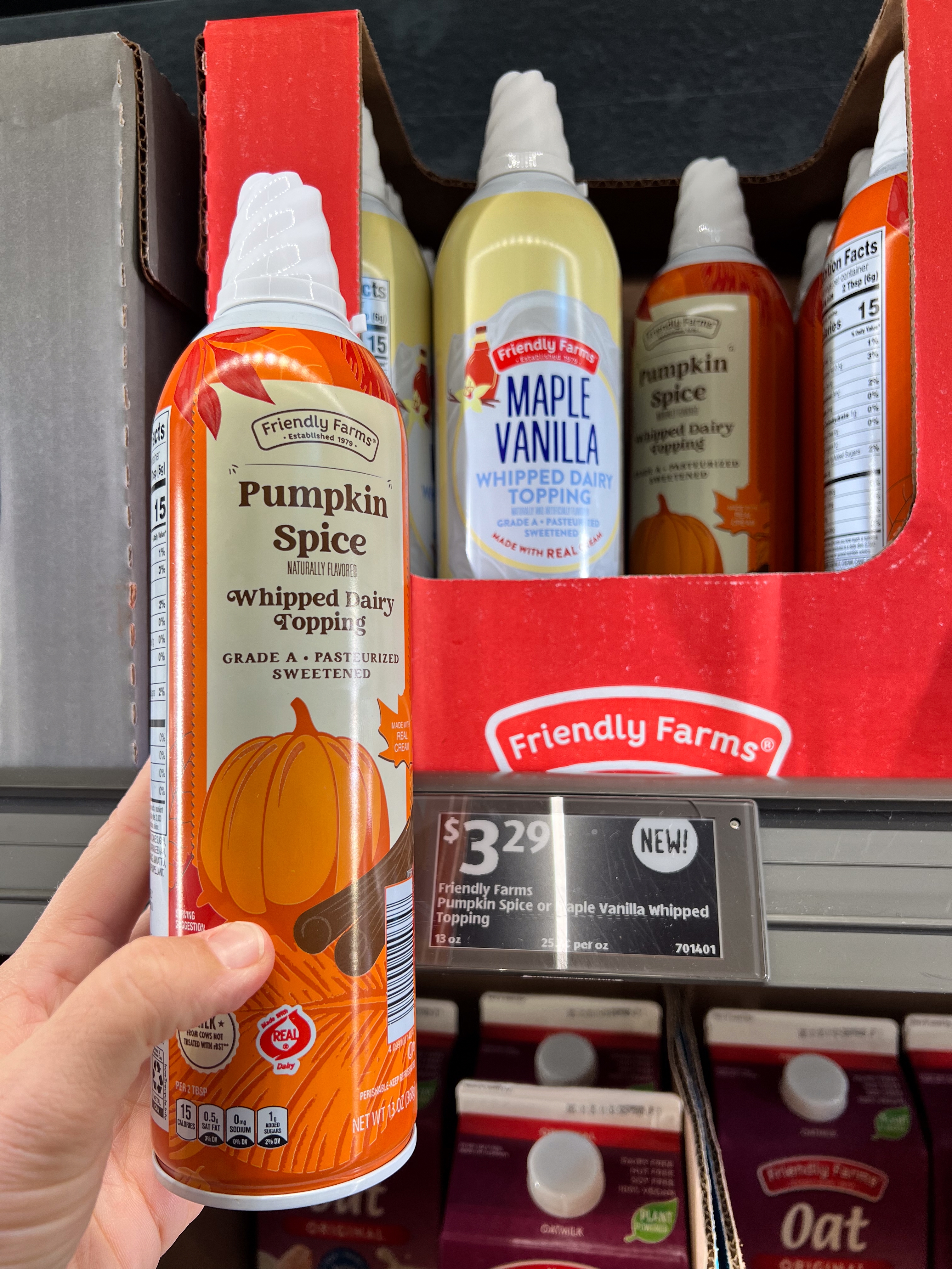 Hand holding a can of Pumpkin Spice whipped topping in front of a display with other flavors. Price tag reads $3.29