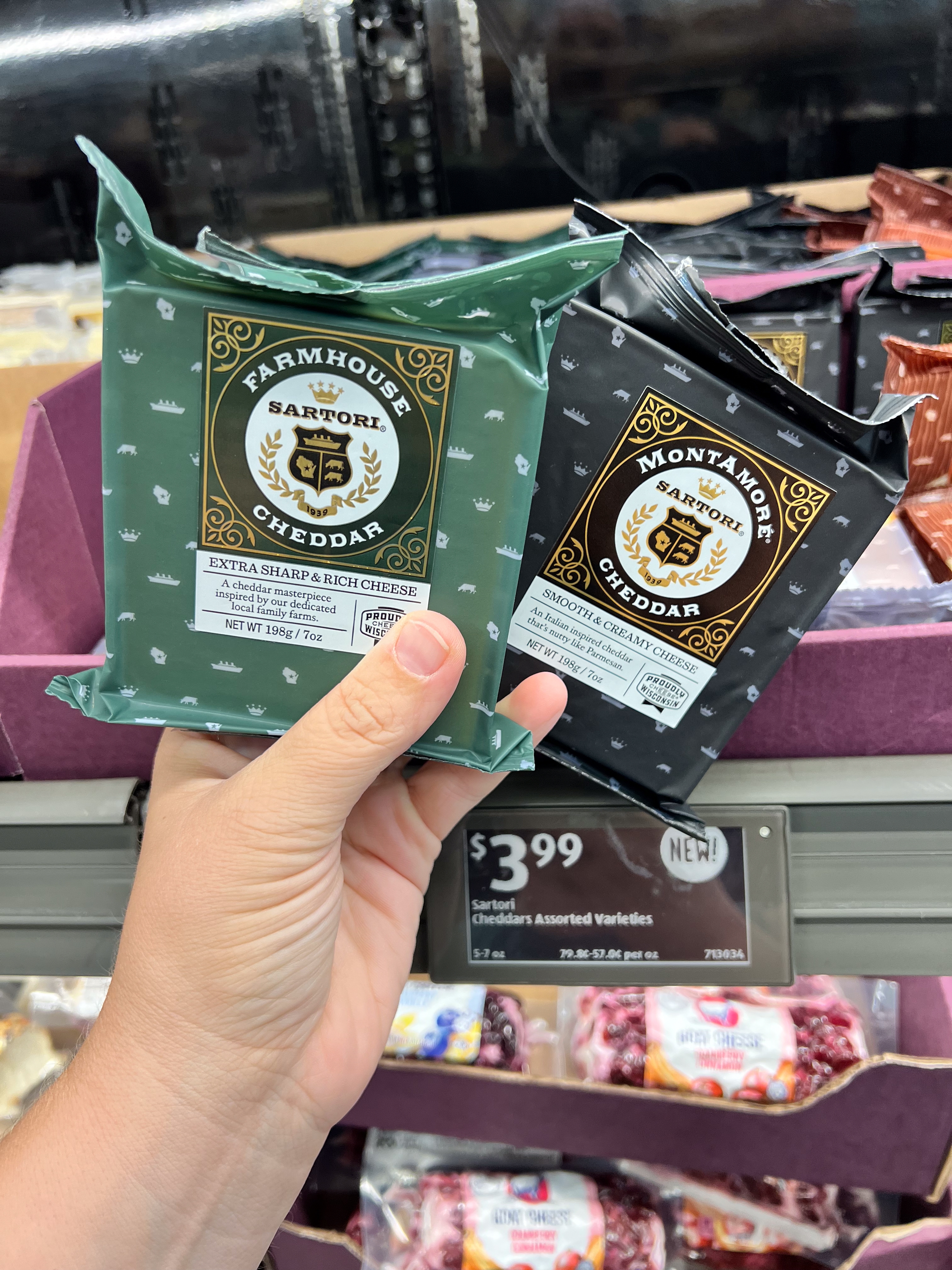 Person holding packs of Sartori Farmhouse Cheddar and MontAmoré Cheddar cheese above a grocery shelf