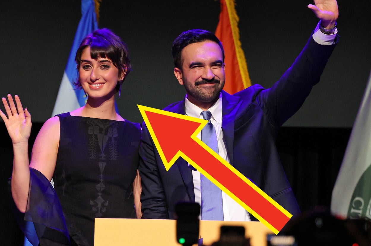 男性と女性が壇上で手を振っている。彼らの後ろには旗が数本立っている。