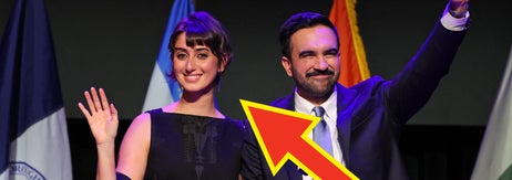 男性と女性が壇上で手を振っている。彼らの後ろには旗が数本立っている。