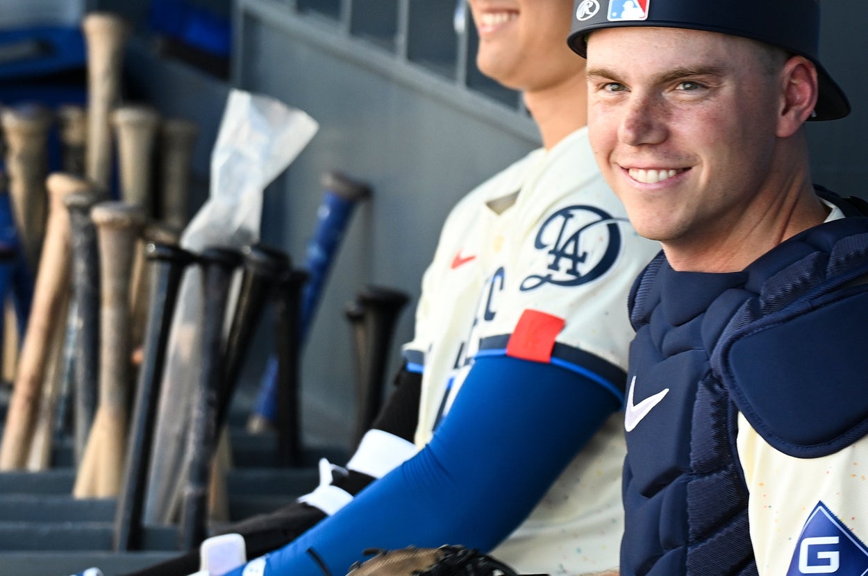 野球選手がベンチでユニフォームとキャッチャー防具を着用し、バットラックの前でリラックスして座っている。