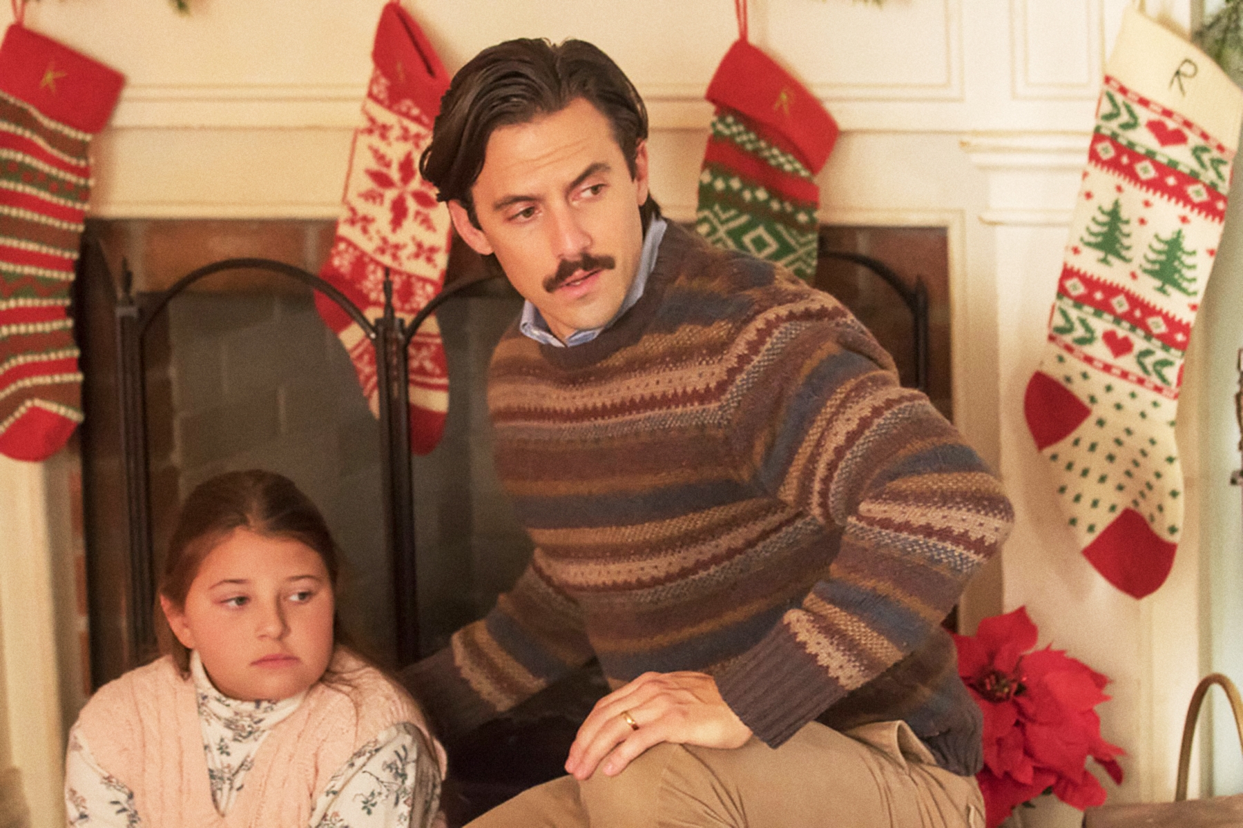 Man with a mustache successful  a striped sweater sits beside a young miss  successful  beforehand   of a fireplace decorated with Christmas stockings