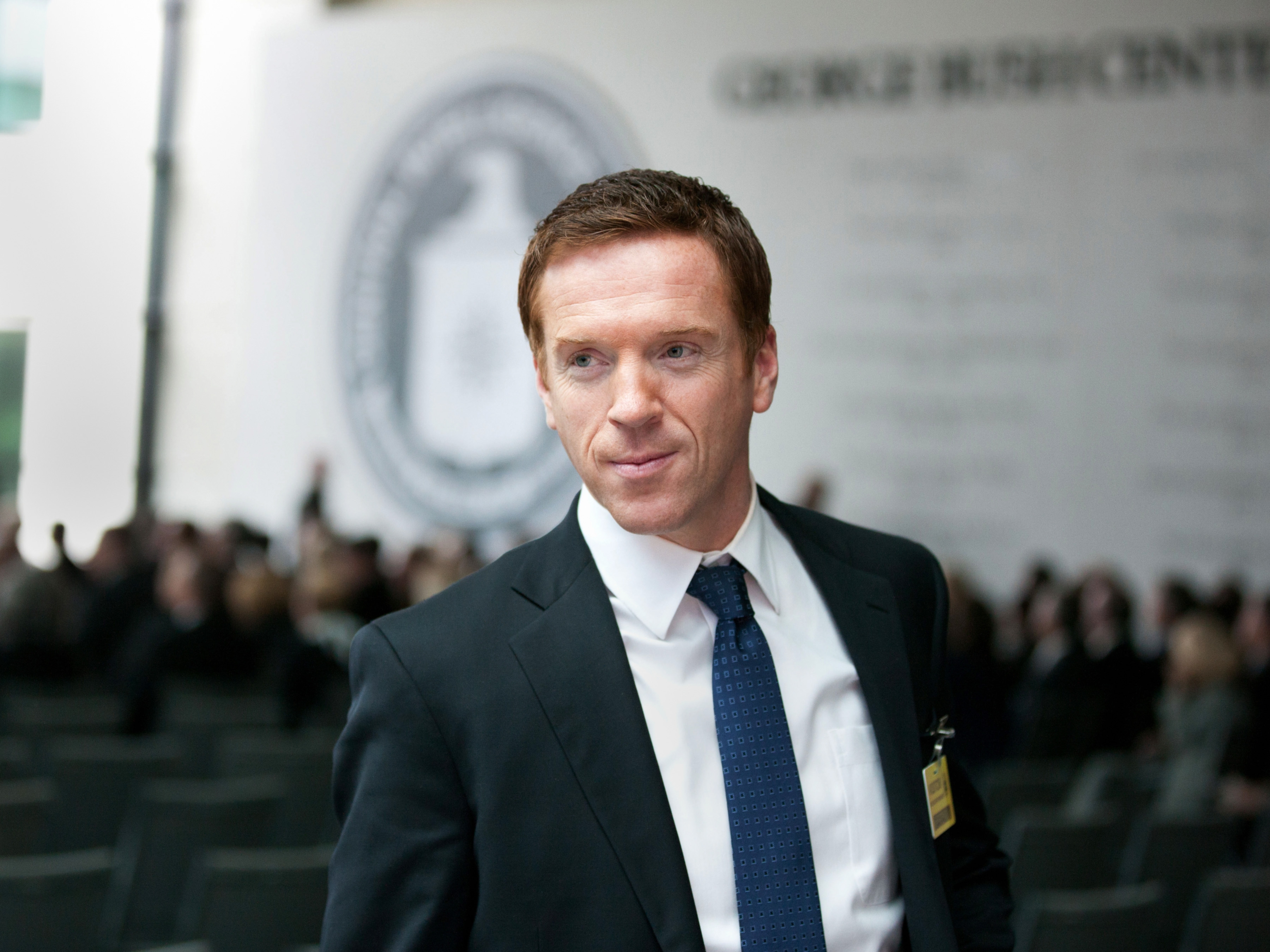 A idiosyncratic   successful  a suit   and necktie  stands successful  a ceremonial  setting, with a blurred assemblage  and a ample  emblem successful  the background