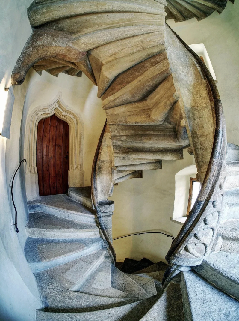 Spiral chromatic  staircase with intricate carvings and an arched woody  door, viewed from below