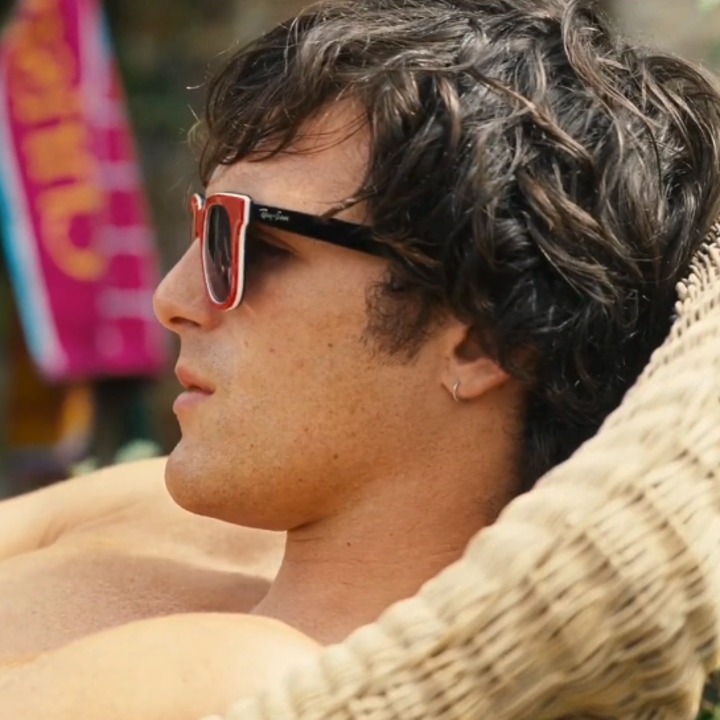 Person wearing sunglasses relaxes on a wicker chair outdoors, with a towel in the background