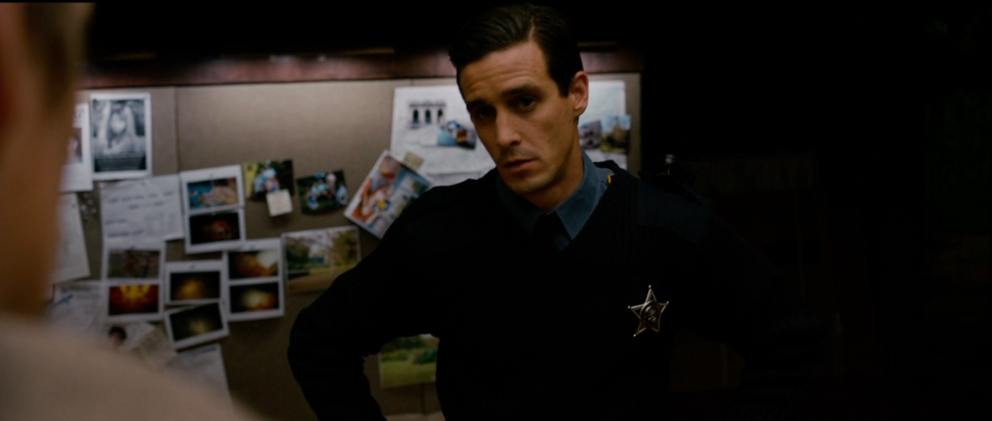 Person successful  a constabulary  azygous   with a badge stands successful  beforehand   of a partition  covered with photos and notes, appearing superior   and engaged successful  conversation