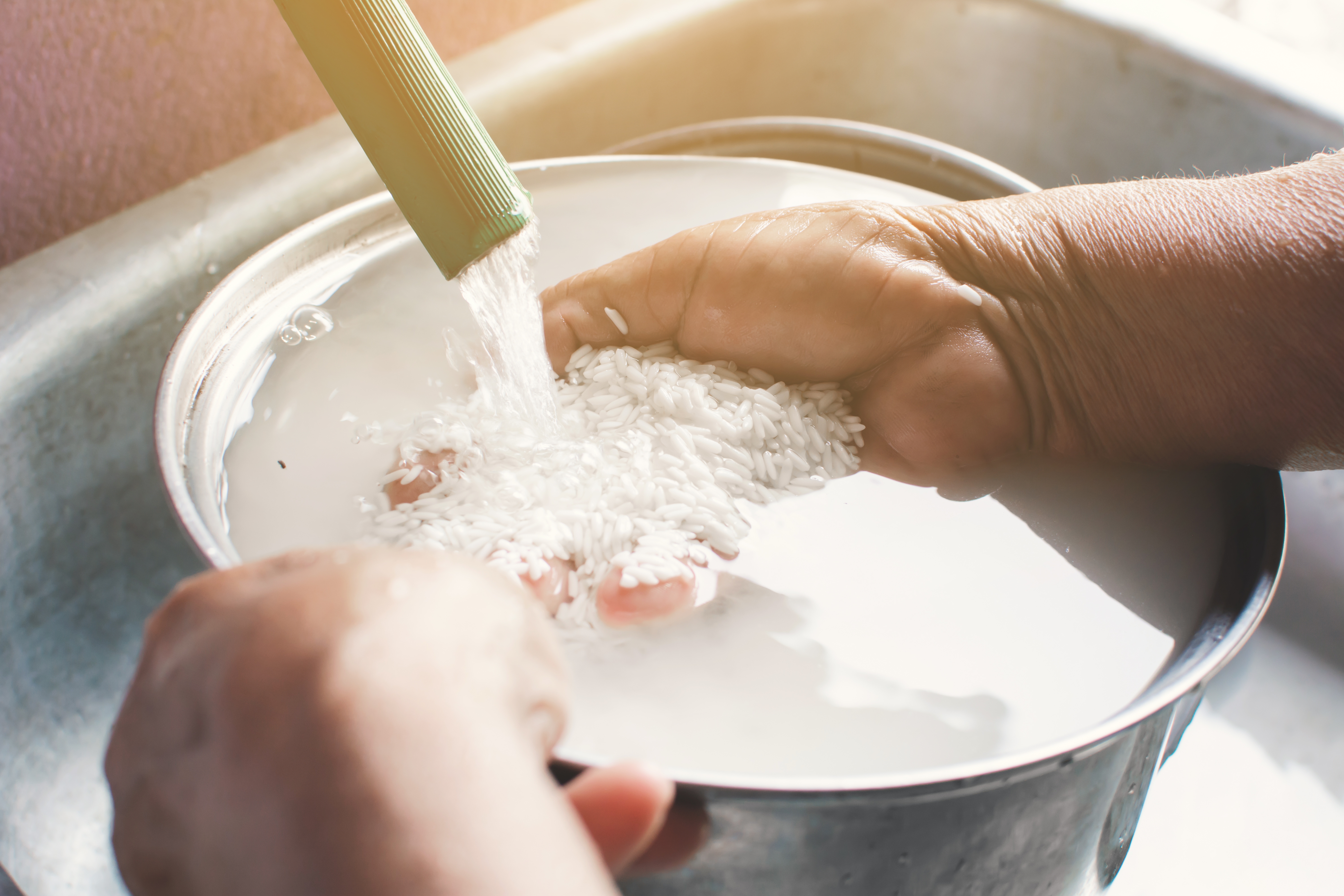 Hands rinsing rice under running water in a metal bowl in a kitchen setting