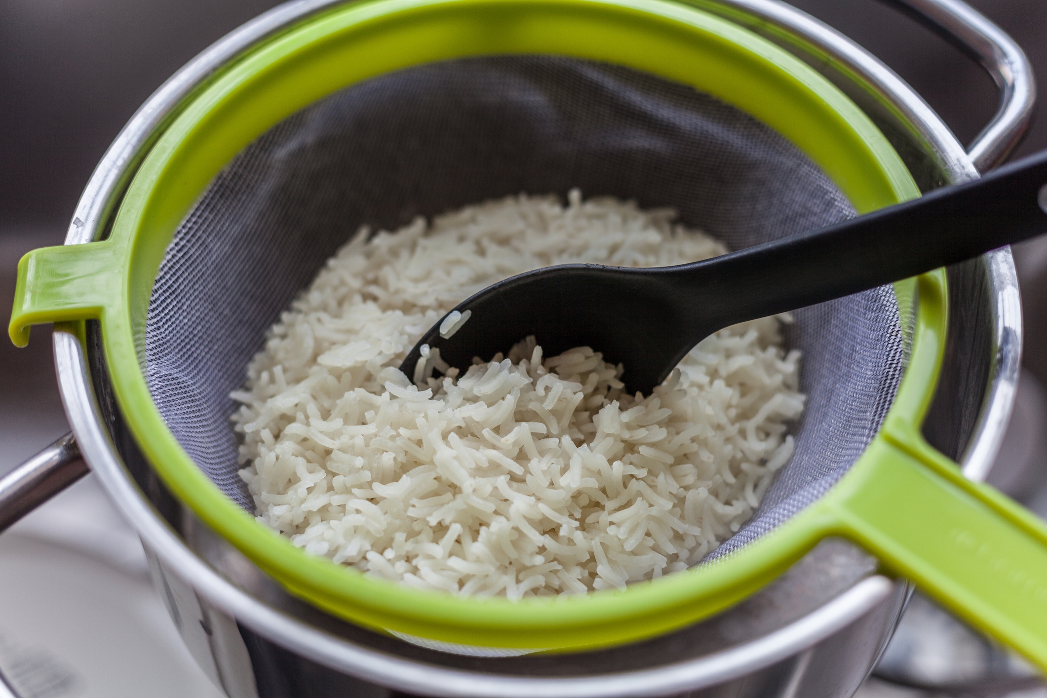 Cooked rice being drained in a mesh strainer placed over a pot, with a black spoon resting on the rice