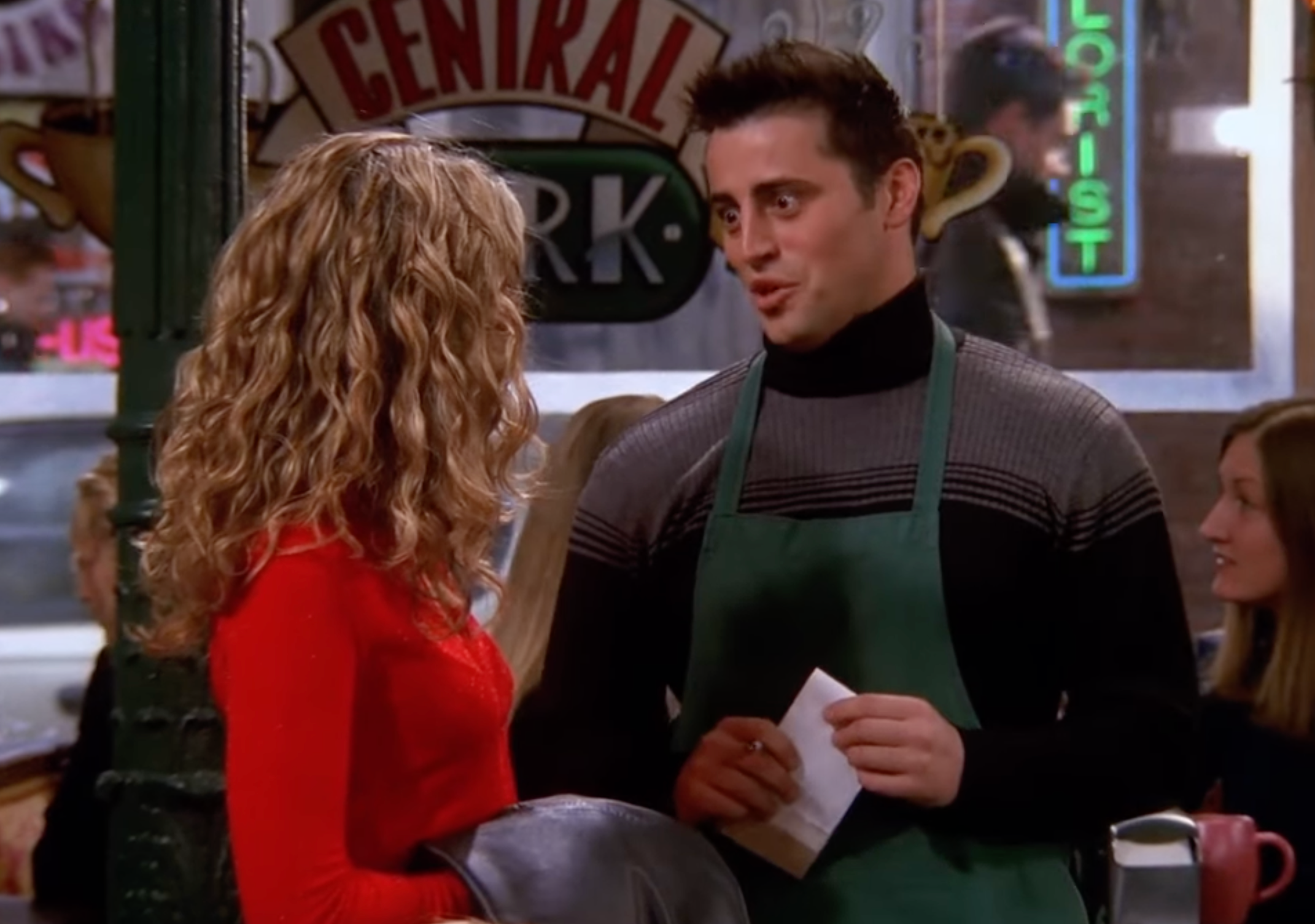 Two people are conversing in a café setting. One wears a turtleneck and apron, holding a notepad. The other has curly hair, dressed casually