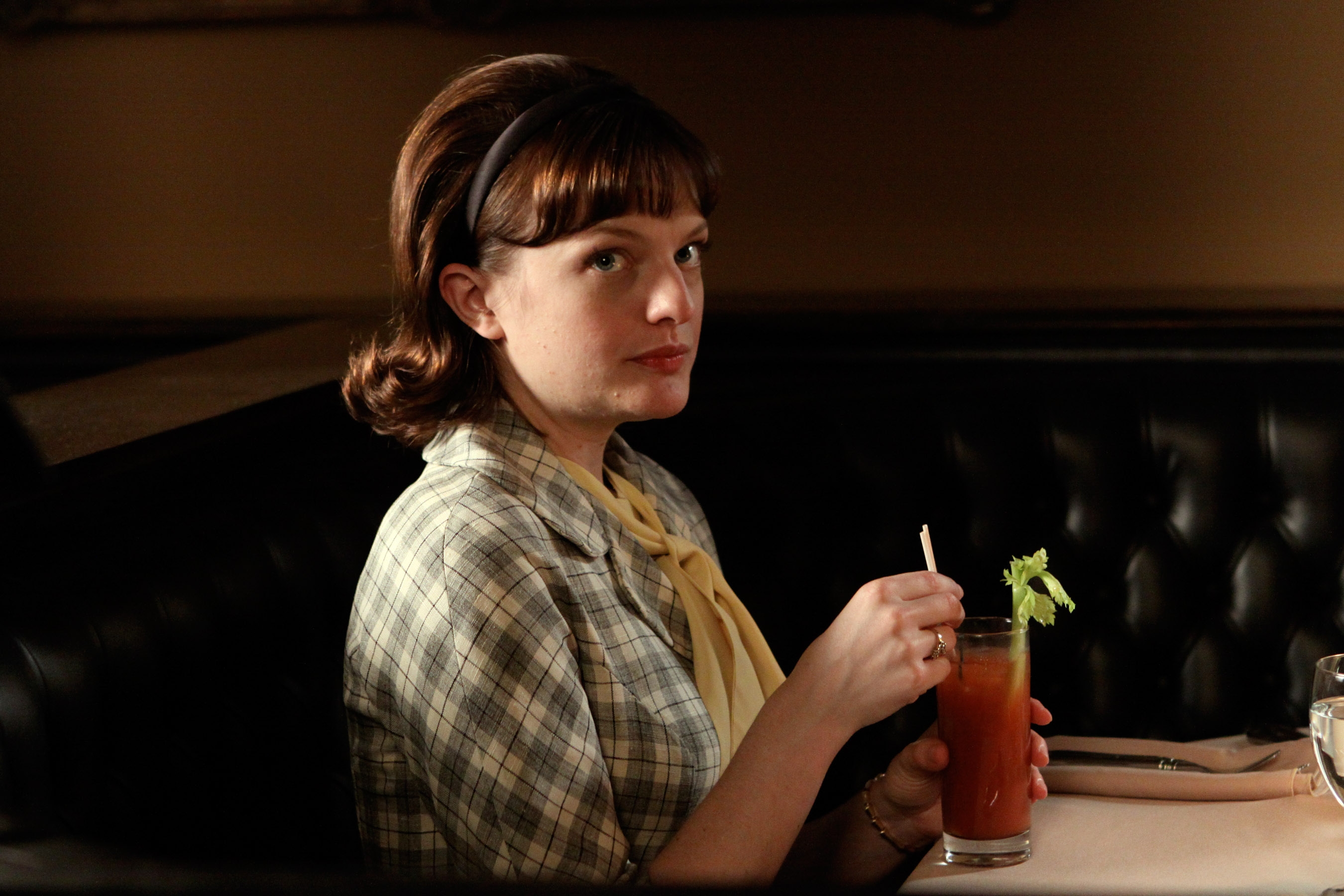 Person in a checkered jacket, sipping a drink with a celery garnish, seated in a restaurant booth