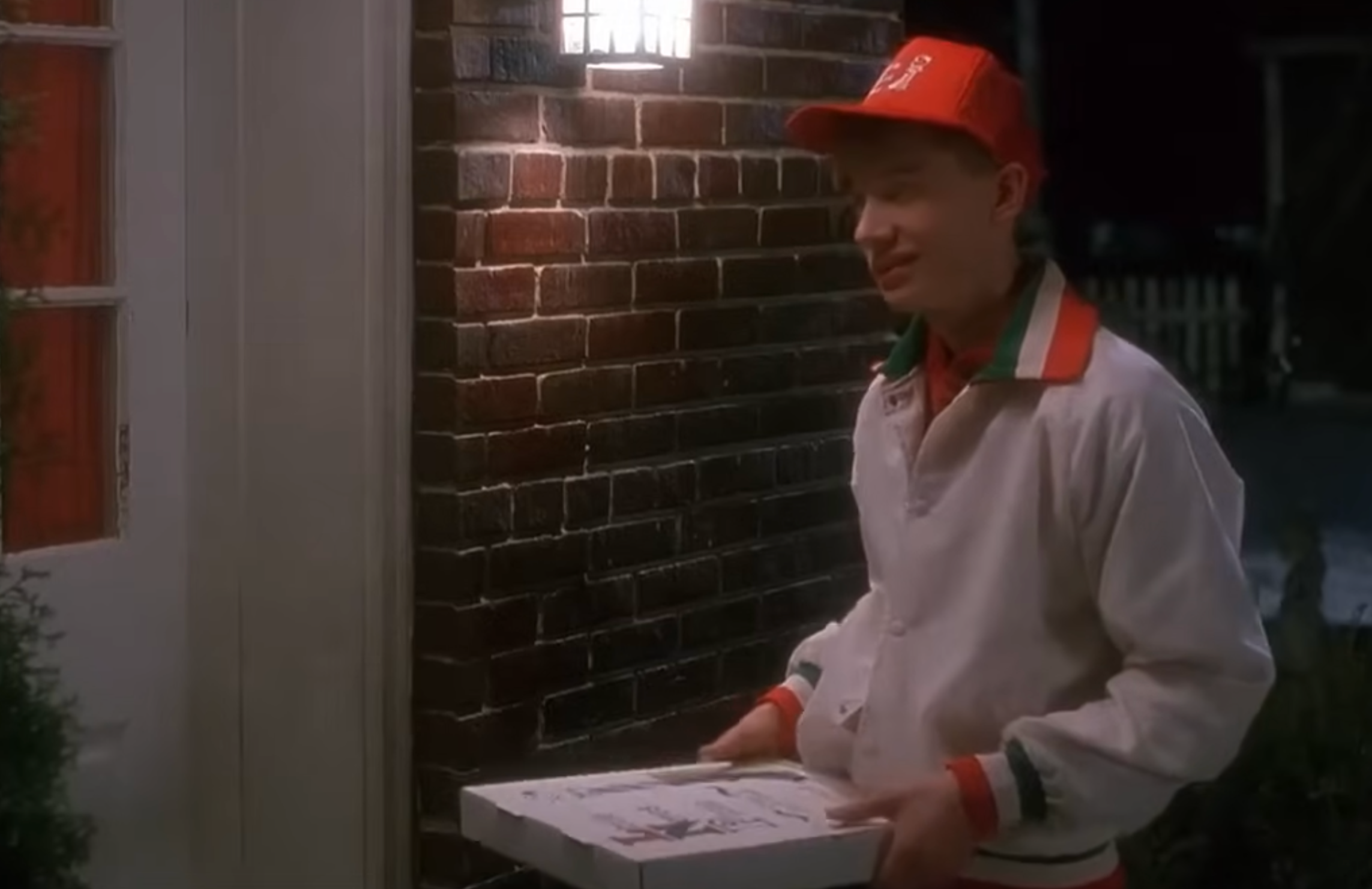 Person in delivery uniform holding pizza box outside a house at night, suggesting a home delivery service
