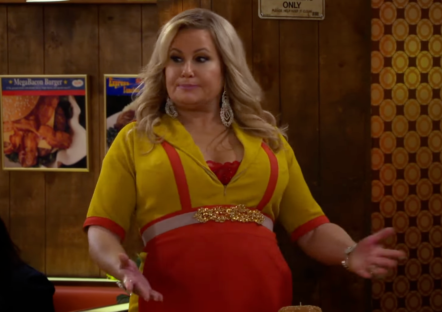 A woman in a diner, wearing a red and yellow outfit with ornate earrings, gestures with her hands. Wall decor featuring food is visible in the background