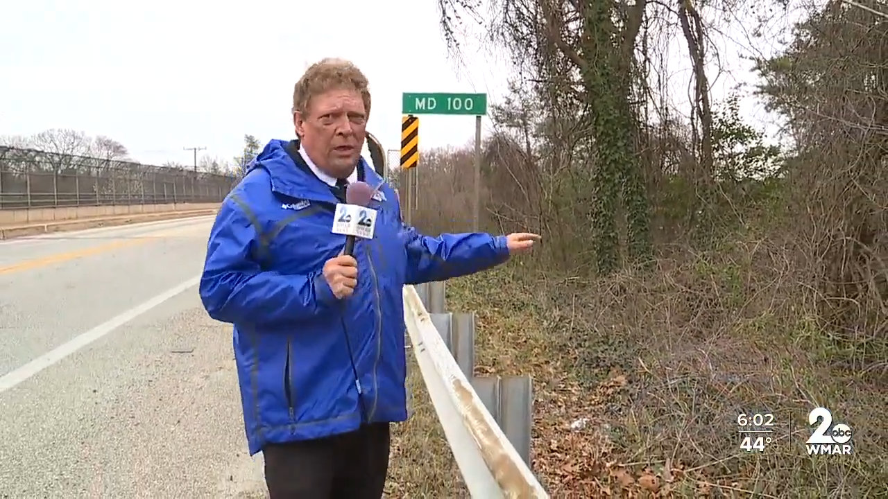 Reporter stands by roadworthy obstruction adjacent MD 100 sign, gesturing towards wooded area