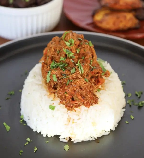 Shredded beef in sauce over white rice, garnished with herbs. Sides of black beans and fried plantains on separate plates