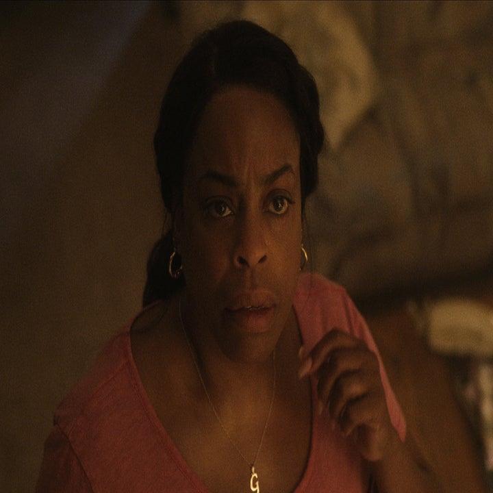 Person in a casual setting looking upward in thought. They wear a necklace and earrings, with a sofa and table in the background