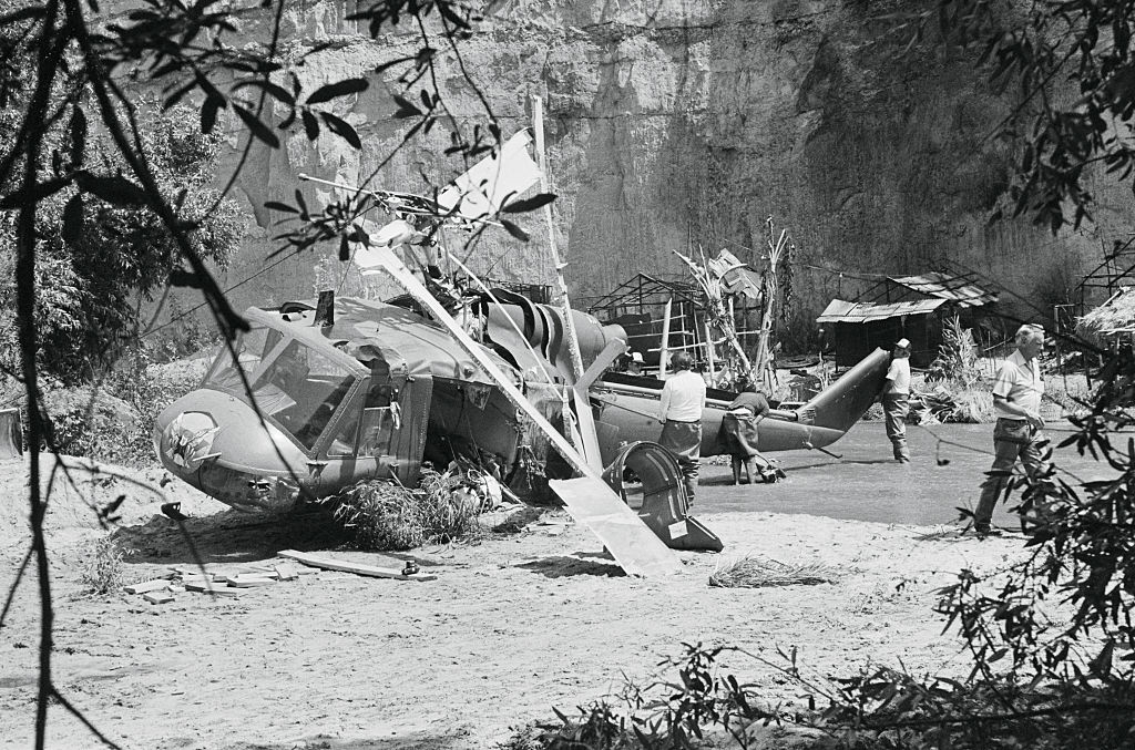 Helicopter clang landing country from the filming of a movie, with acceptable workers examining the area, surrounded by makeshift huts and foliage