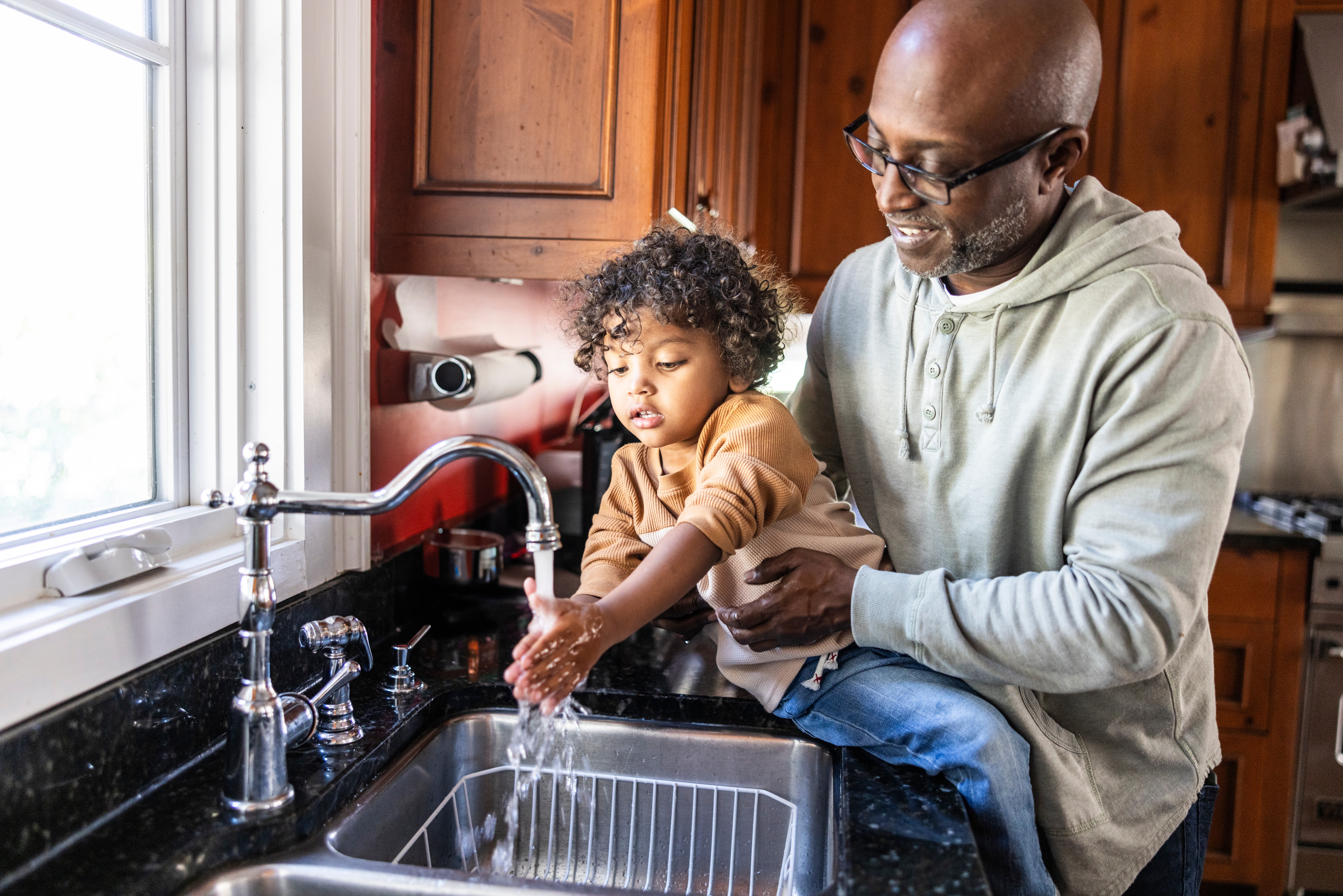 Foreldre hjelper småbarn med å vaske hender ved kjøkkenvasken, både smilende og nyter øyeblikket. Varm familieinteraksjon i koselige hjemlige omgivelser