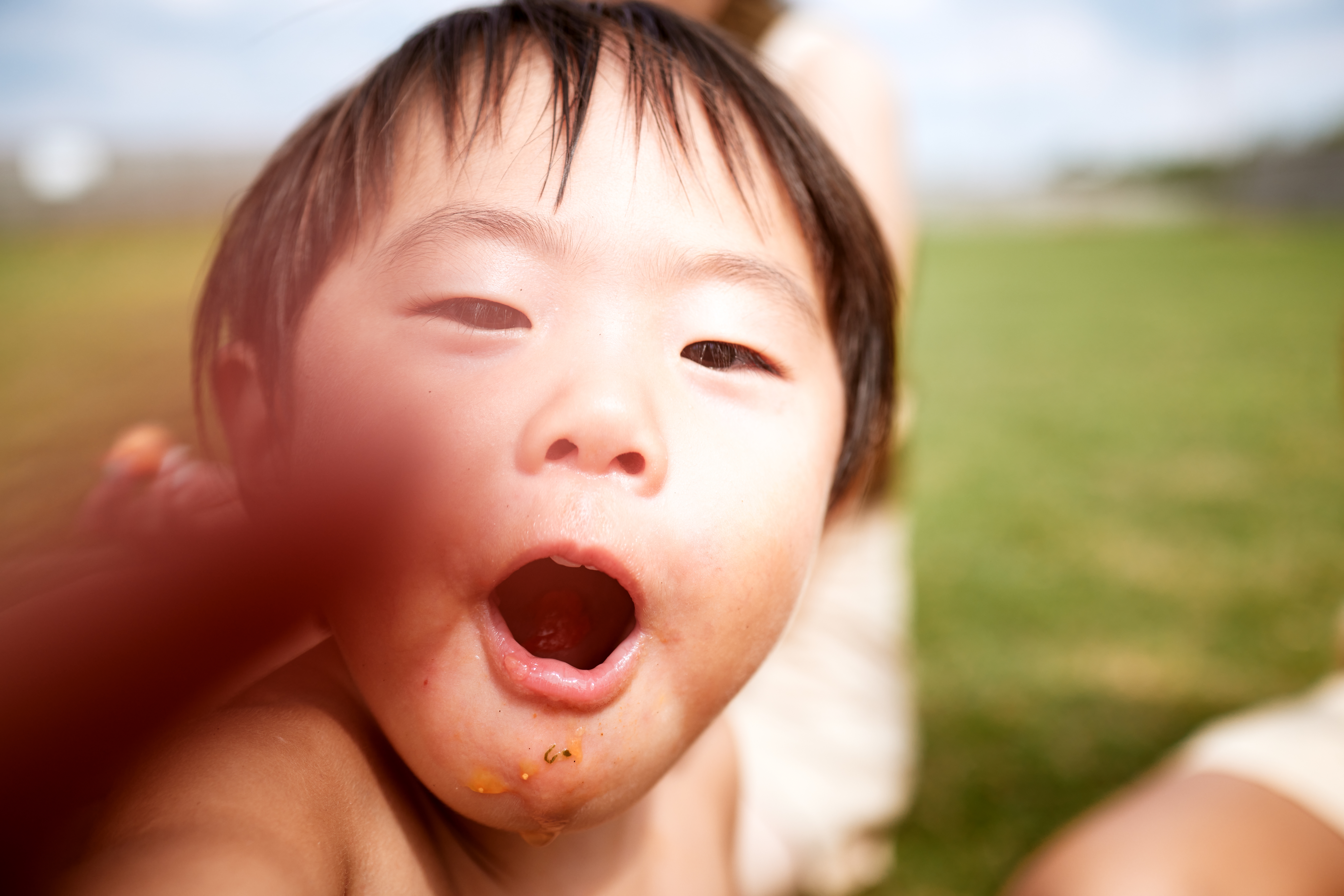 Nærbilde av et barn utendørs med åpen munn og lekent uttrykk, fingrene som delvis dekker linsen