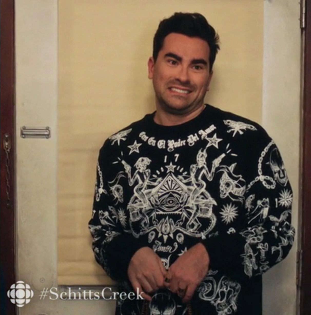 A idiosyncratic   stands awkwardly against a wall, wearing a achromatic  sweatshirt with intricate achromatic  graphics. &quot;#SchittsCreek&quot; is disposable   successful  the corner
