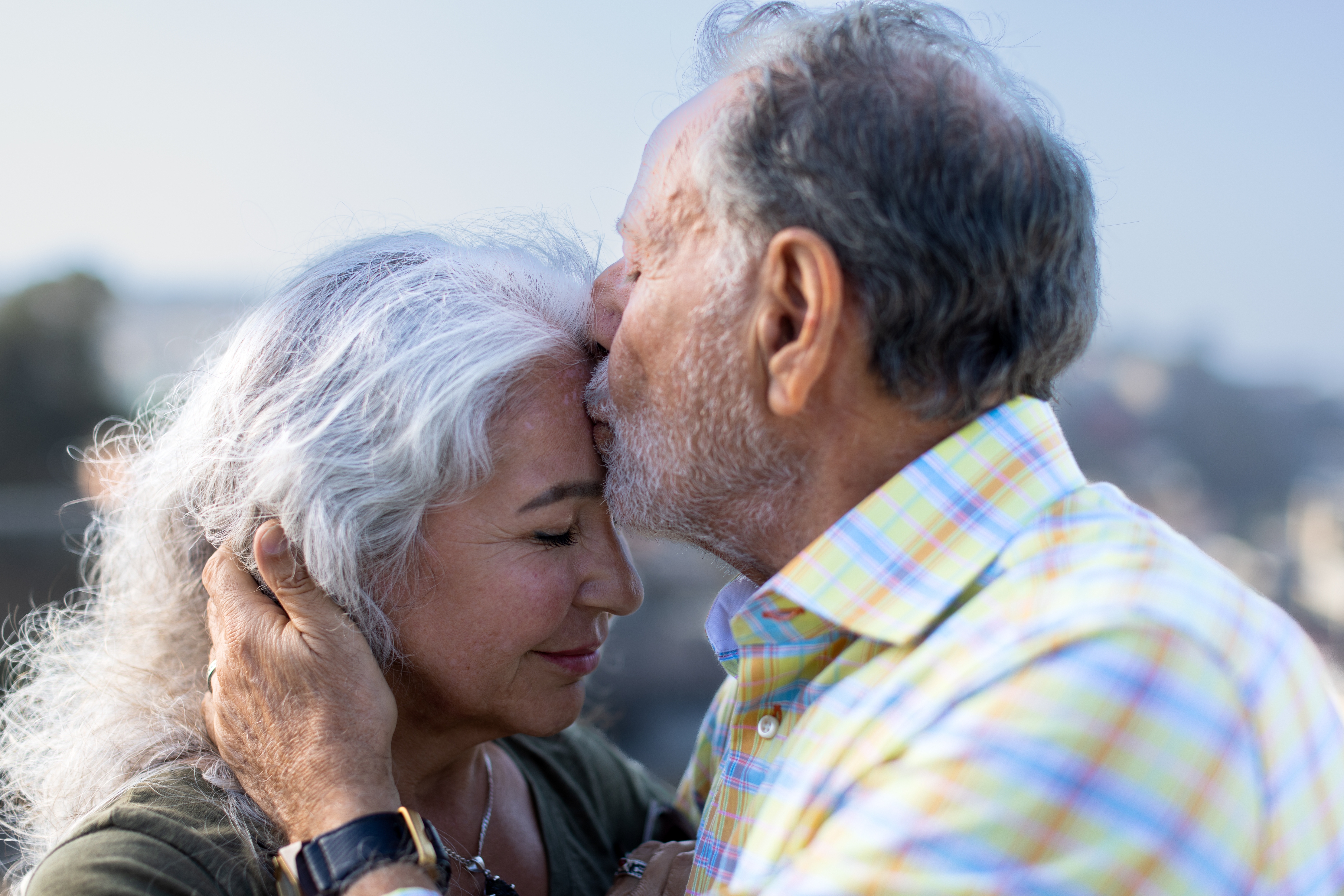 Eldre mann kysser forsiktig en eldre kvinnes panne utendørs, begge viser øm hengivenhet
