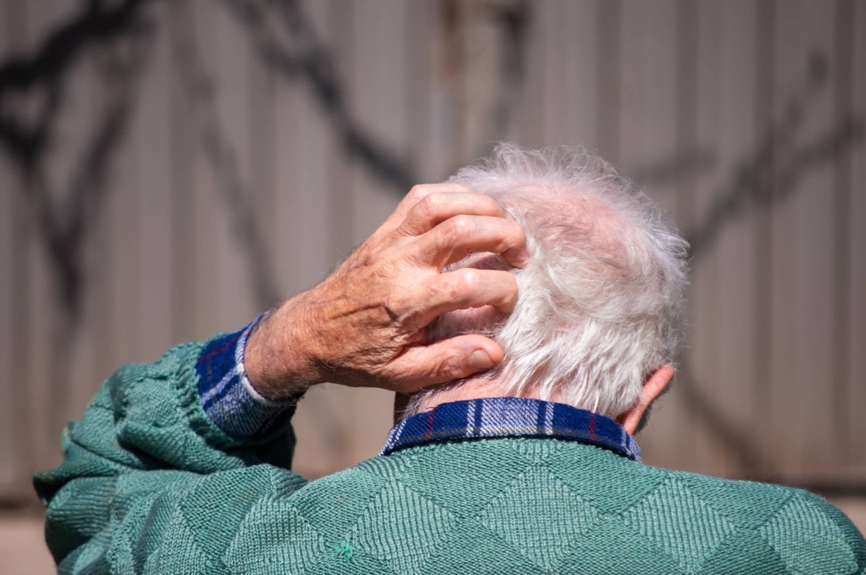 An elderly person scratches their head while facing away, wearing a textured sweater over a plaid-collared shirt, standing outside