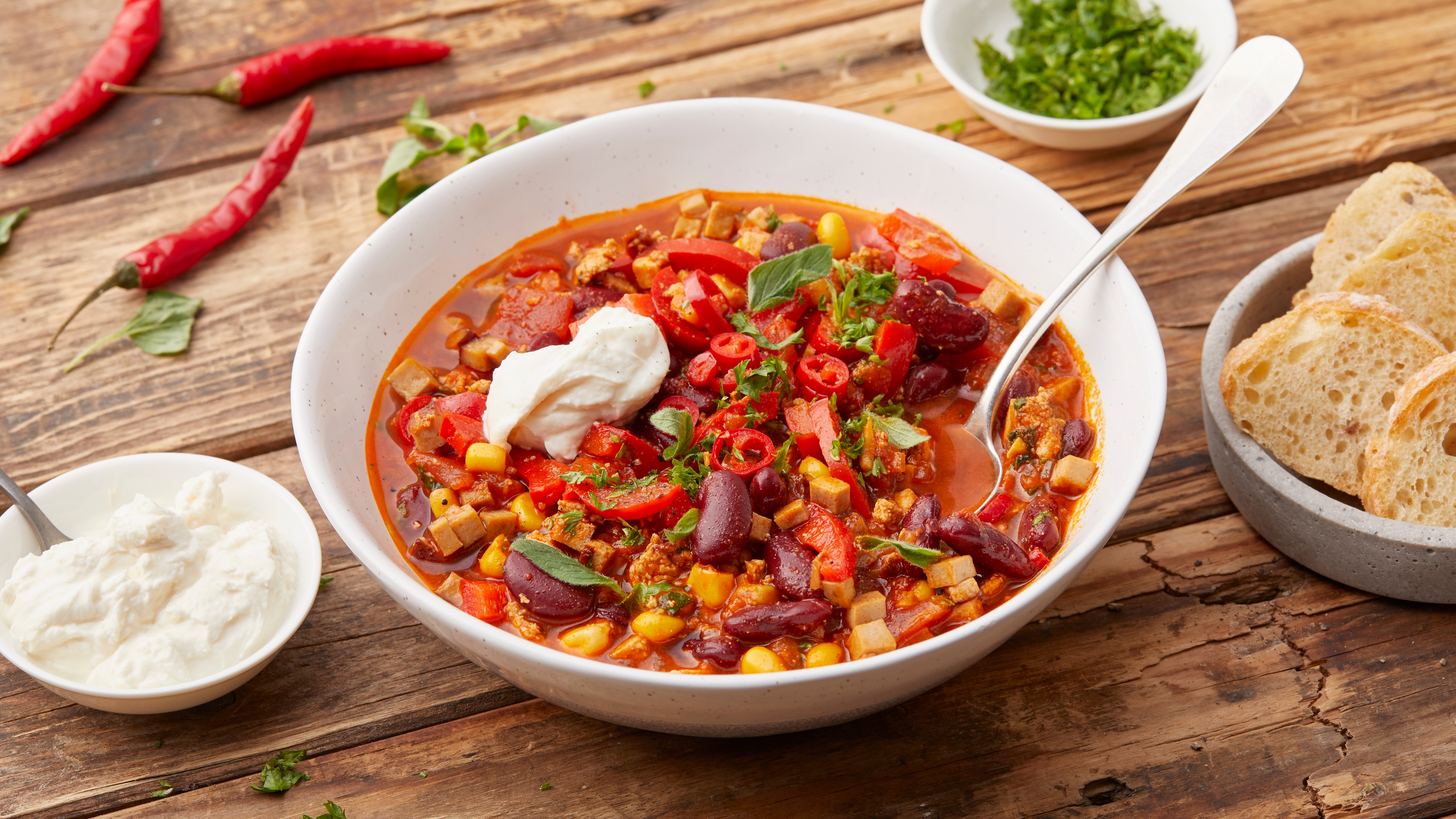 Bowl of chili with toppings, surrounded by bread and seasoning dishes on a wooden table