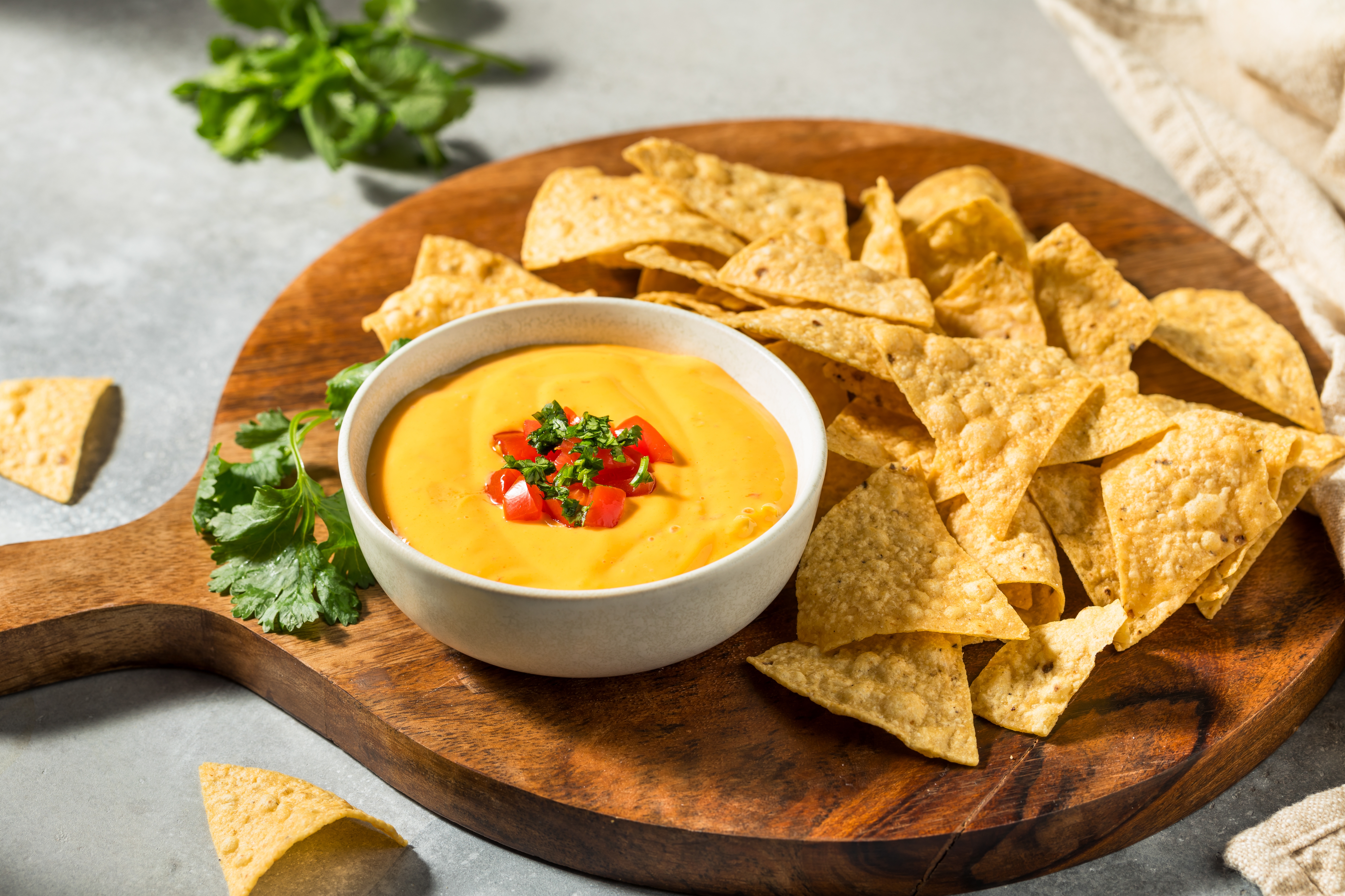 Tortilla chips surround a bowl of cheese dip topped with diced tomatoes and herbs on a wooden board