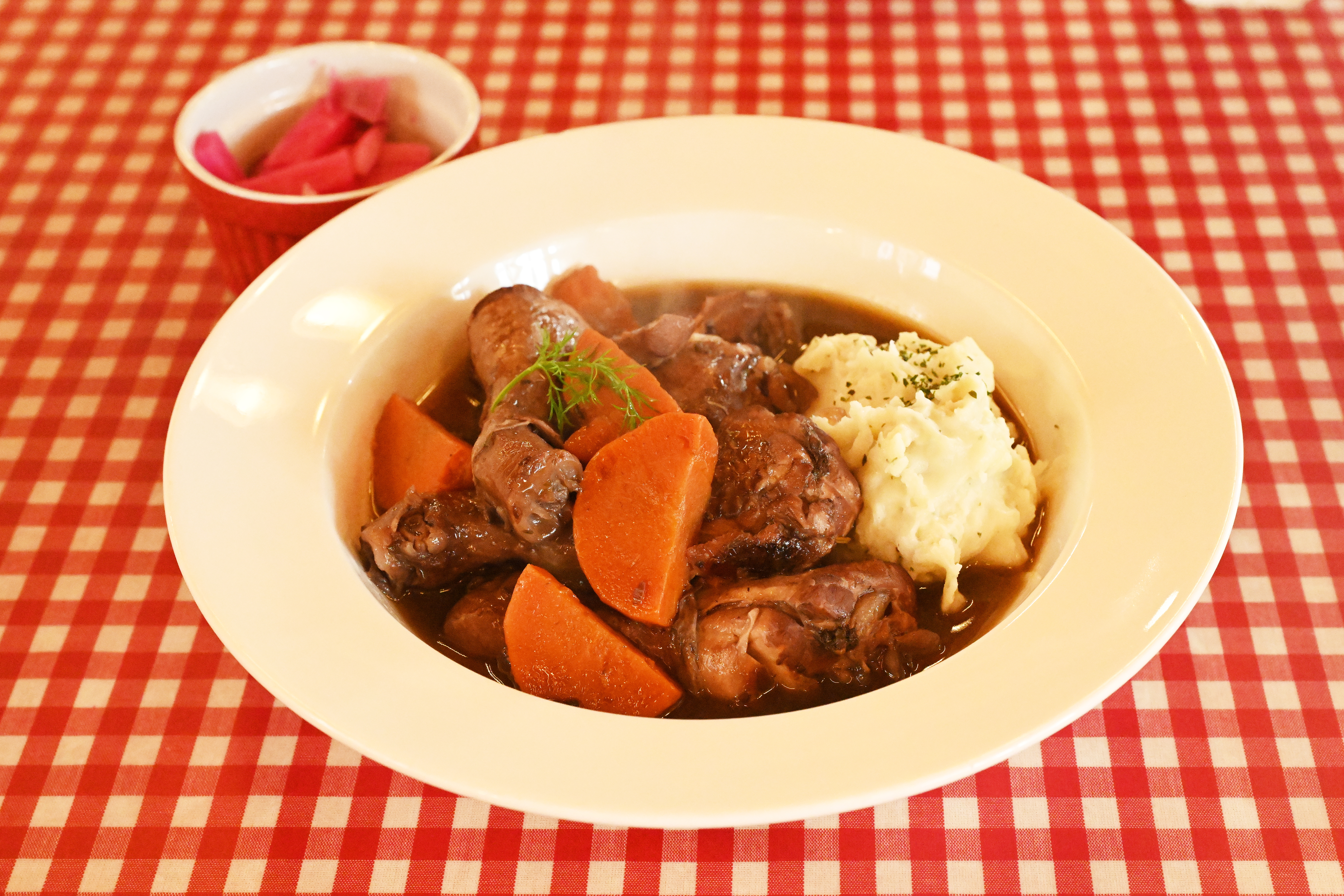 A bowl of stew with carrots, meat, and mashed potatoes on a checkered tablecloth, accompanied by a side dish in a small bowl
