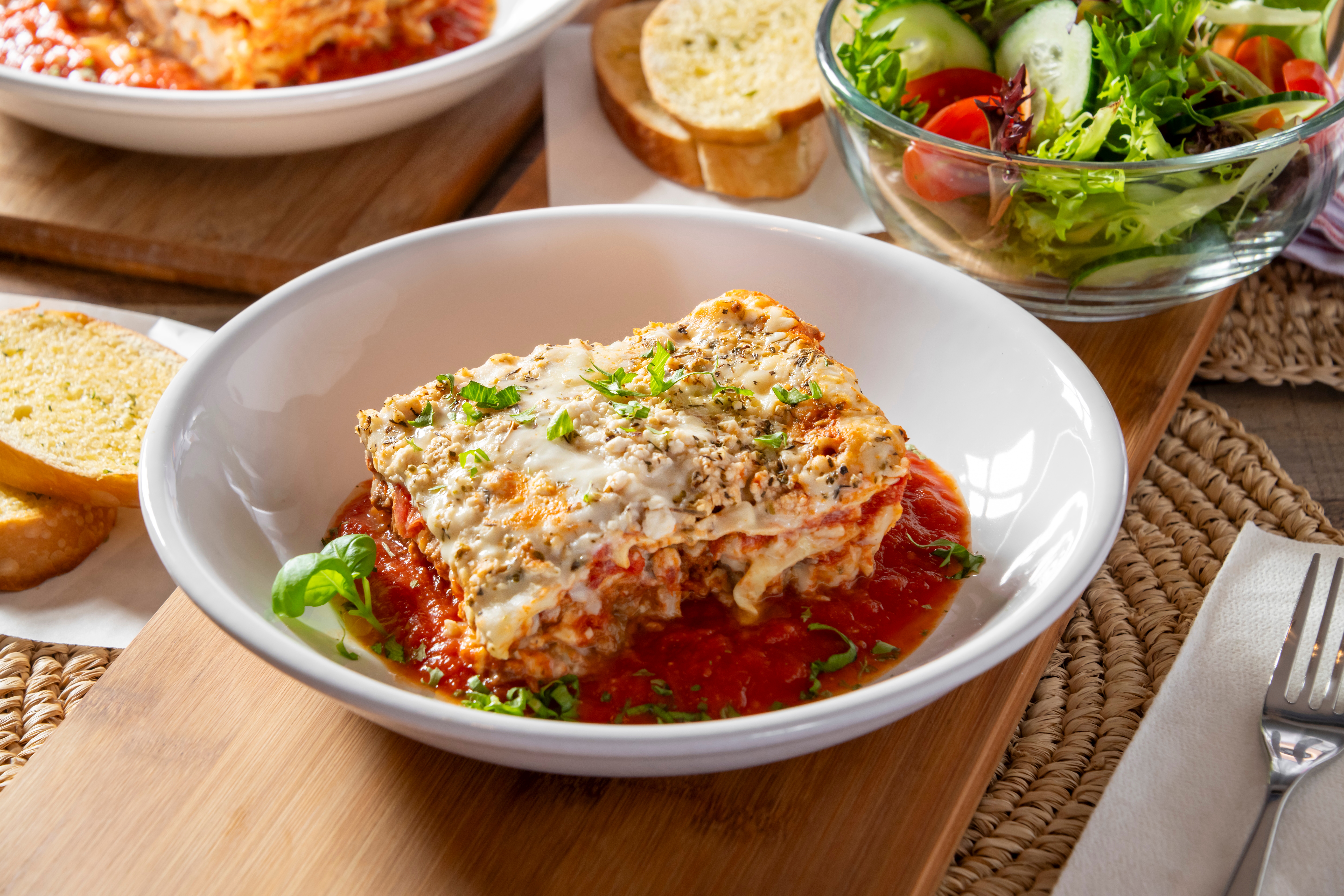 Lasagna served with tomato sauce and herbs, accompanied by garlic bread and a fresh salad with cucumbers and tomatoes