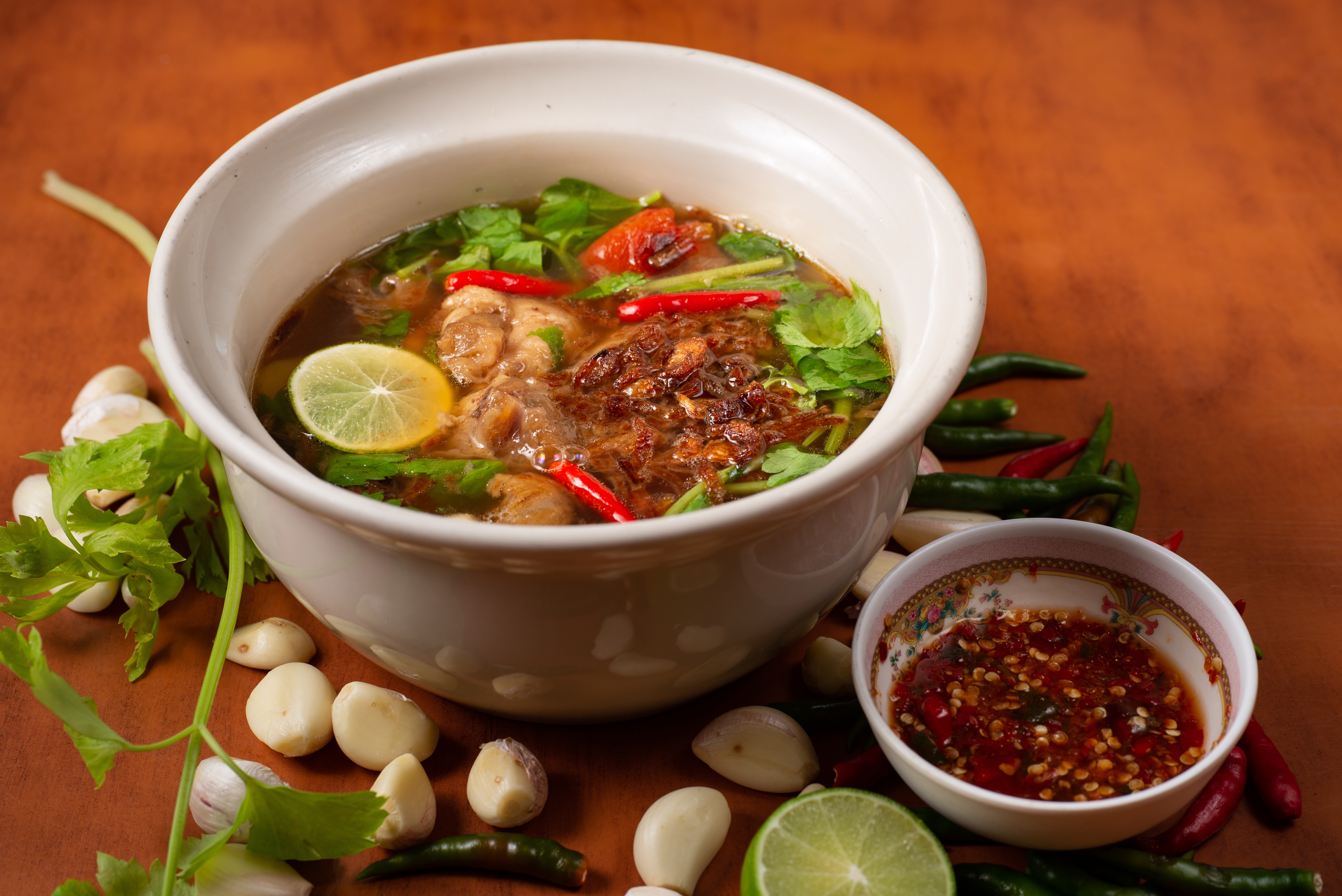 A bowl of soup with meat and herbs, accompanied by various herbs and chili peppers, next to a small bowl of dipping sauce